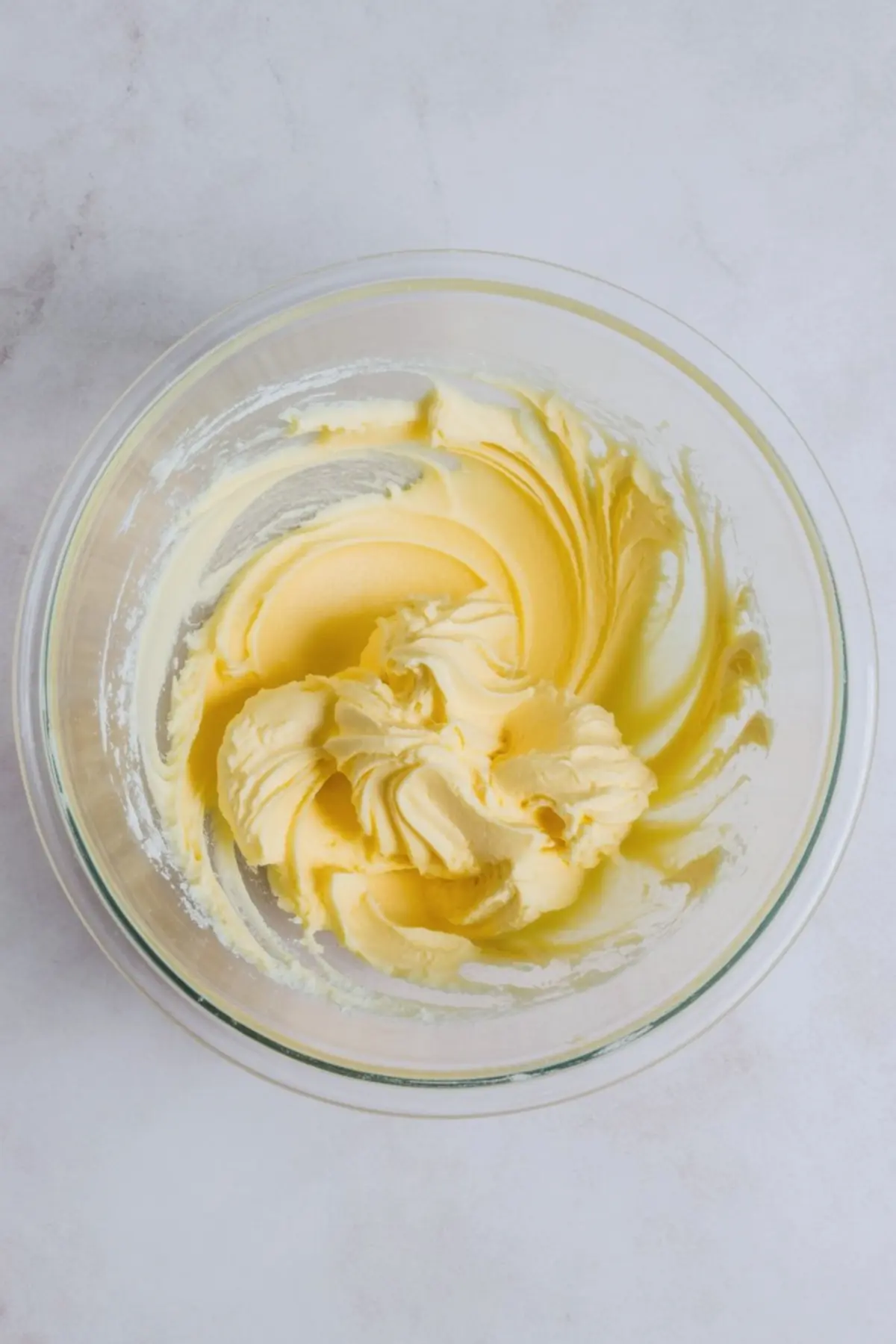Glass mixing bowl holds whipped butter with a smooth, pale yellow texture. Butter shows soft peaks and creamy swirls on a light marble countertop, step of making homemade cookie frosting.
