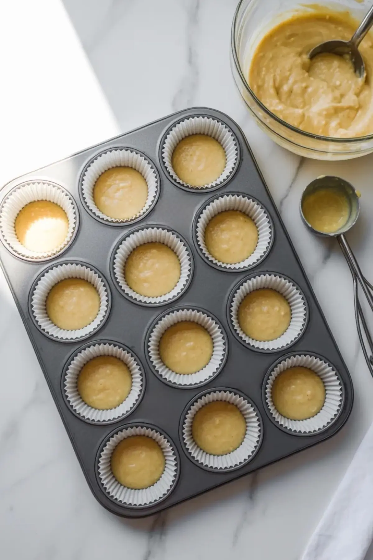 Metal muffin tin holds paper liners filled with cornbread batter on a marble countertop. Glass bowl of batter and measuring scoop sit beside the pan, showing step by step cornbread muffin preparation.
