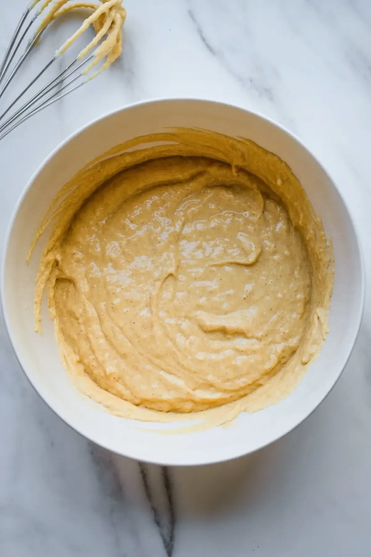 Overhead view shows thick cornbread waffle batter mixed smooth in a white bowl with a whisk coated in batter beside it on a marble countertop, easy homemade waffle recipe texture before cooking.