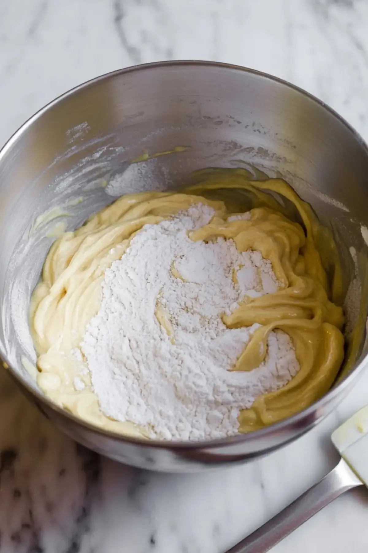Smooth pancake batter in a stainless steel mixing bowl with flour on top, set on a white marble countertop, showing a step in preparing homemade pancake mix.
