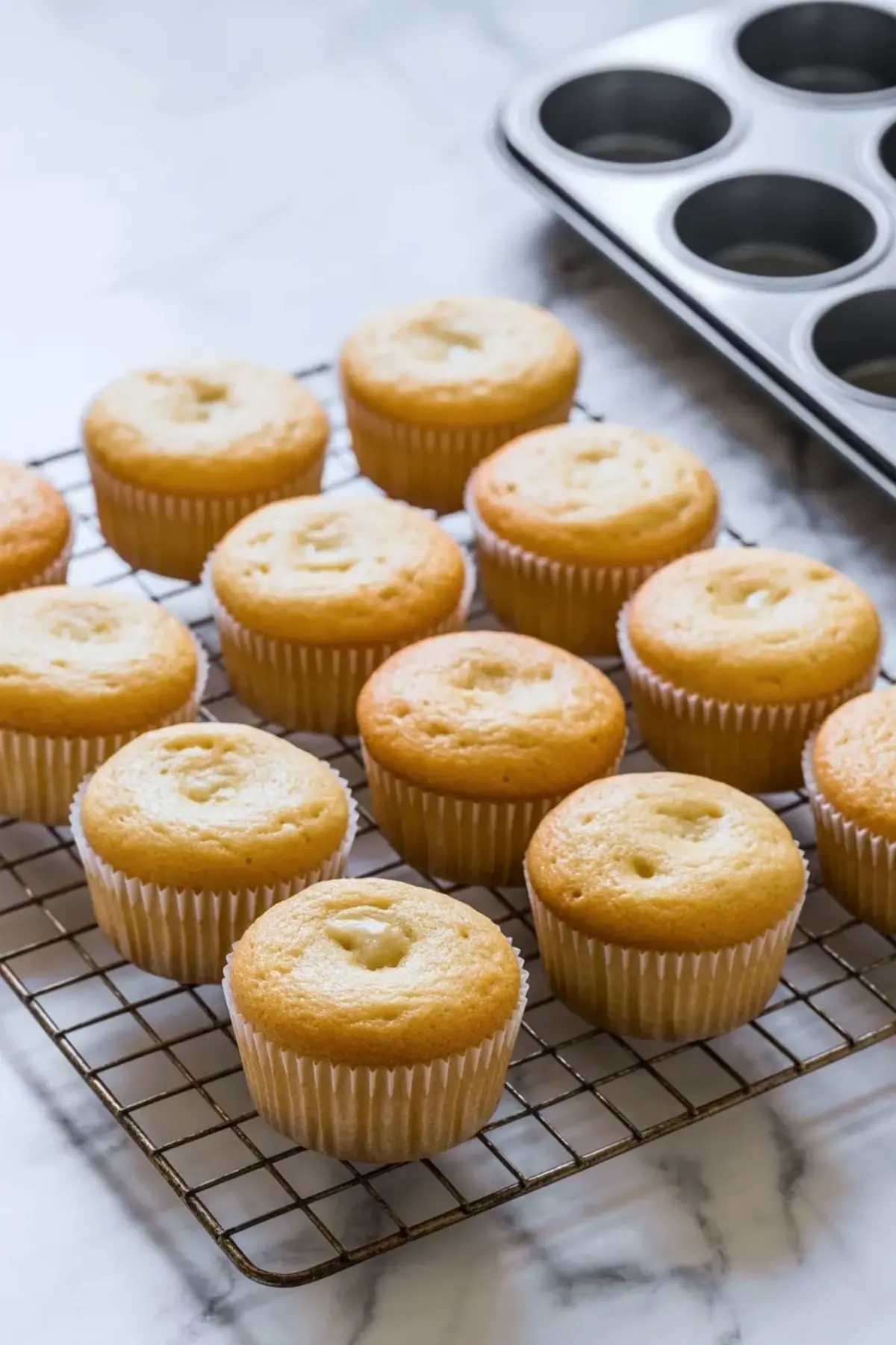 Freshly baked vanilla cupcakes cooling on a wire rack, each with a soft golden top and white cupcake liners, placed beside an empty muffin tin on a marble countertop.
