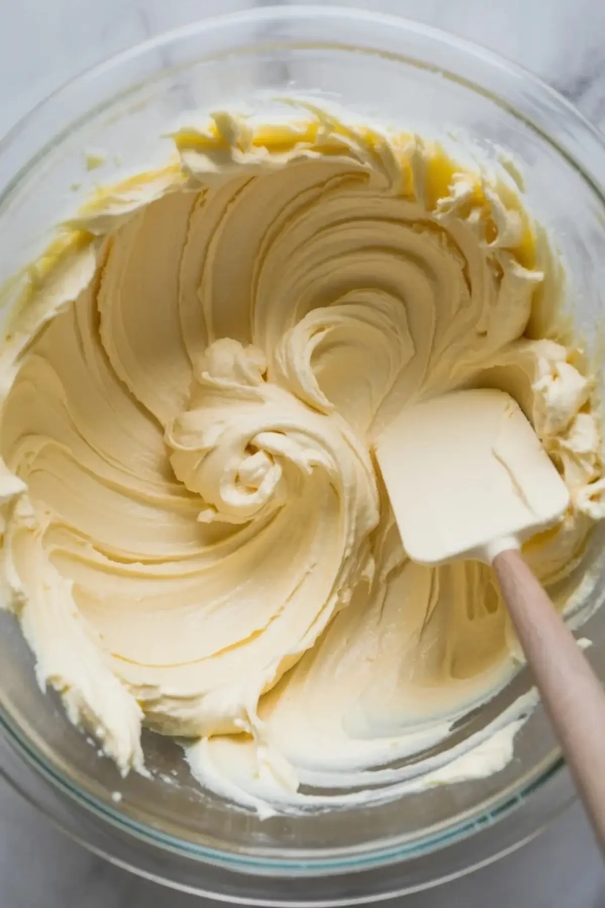 Close-up of thick, fluffy cream cheese filling being swirled with a spatula inside a glass mixing bowl. Homemade frosting or pastry filling with buttery texture.
