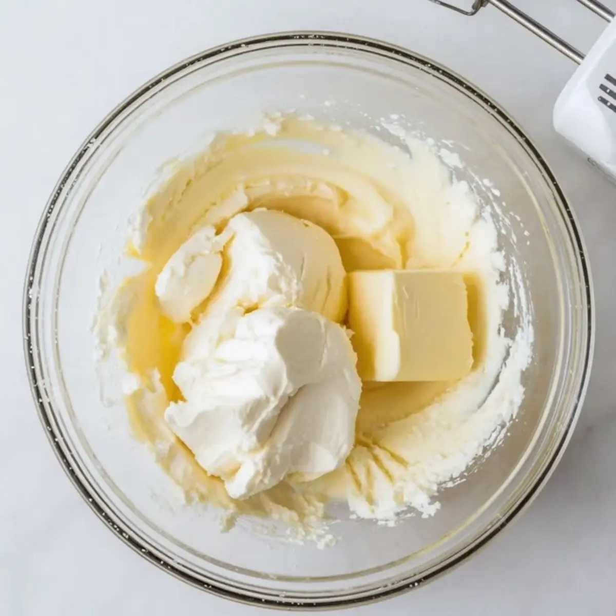 Glass bowl holding a mixture of softened butter and cream cheese, ready to be blended for a dessert filling or frosting. Baking prep image with rich dairy ingredients.
