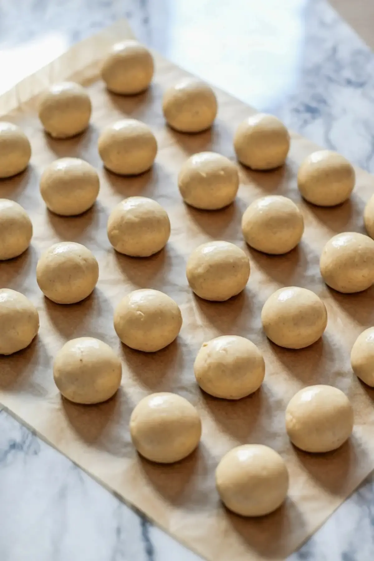 Smooth cookie dough balls rest on parchment paper on a marble countertop, spaced evenly for baking, showing a classic sugar cookie recipe preparation step.
