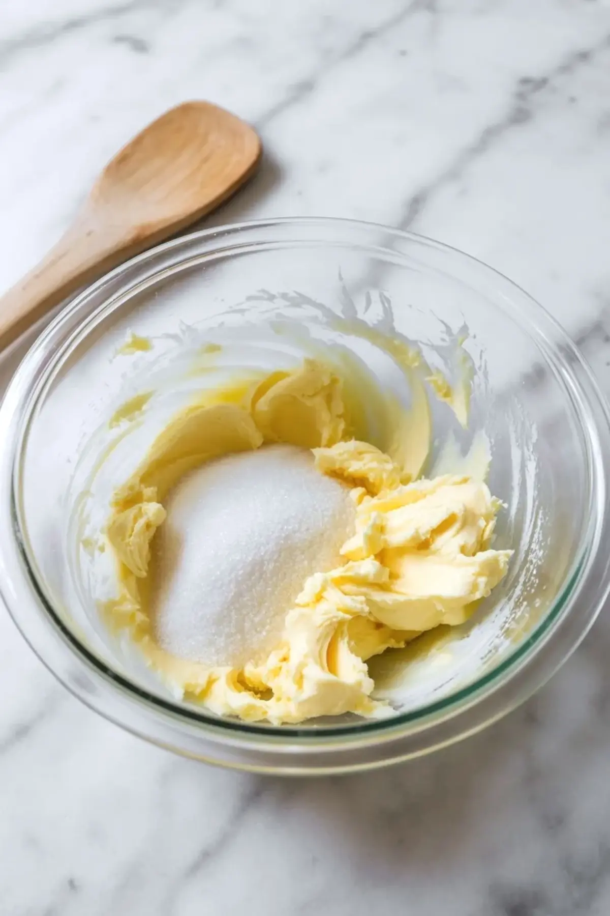 Softened butter and granulated sugar sit in a clear glass mixing bowl with a wooden spoon nearby, showing the creaming step for homemade sugar cookies.
