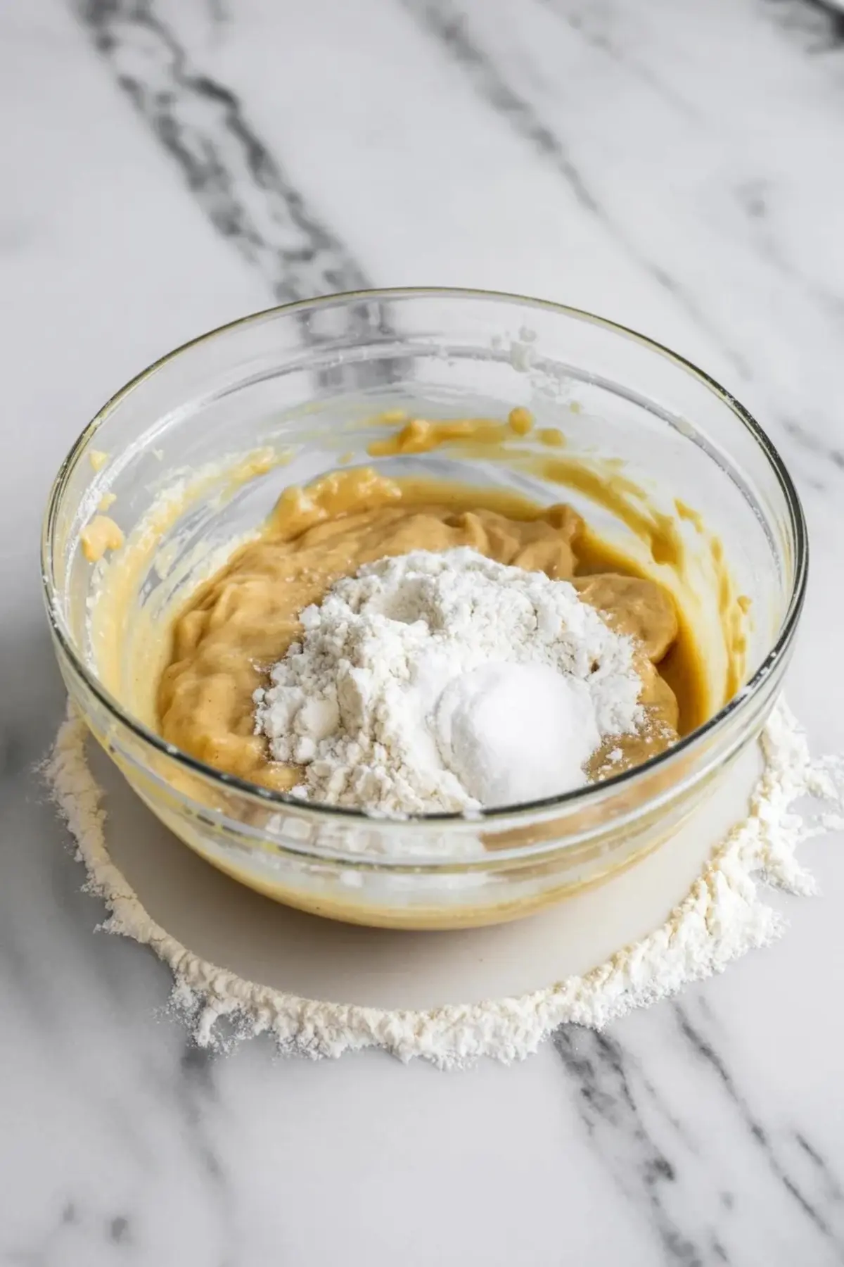 Thick cookie batter holds a mound of flour and sugar in a glass bowl, with flour scattered on the marble surface, showing the dry ingredients added to sugar cookie dough.
