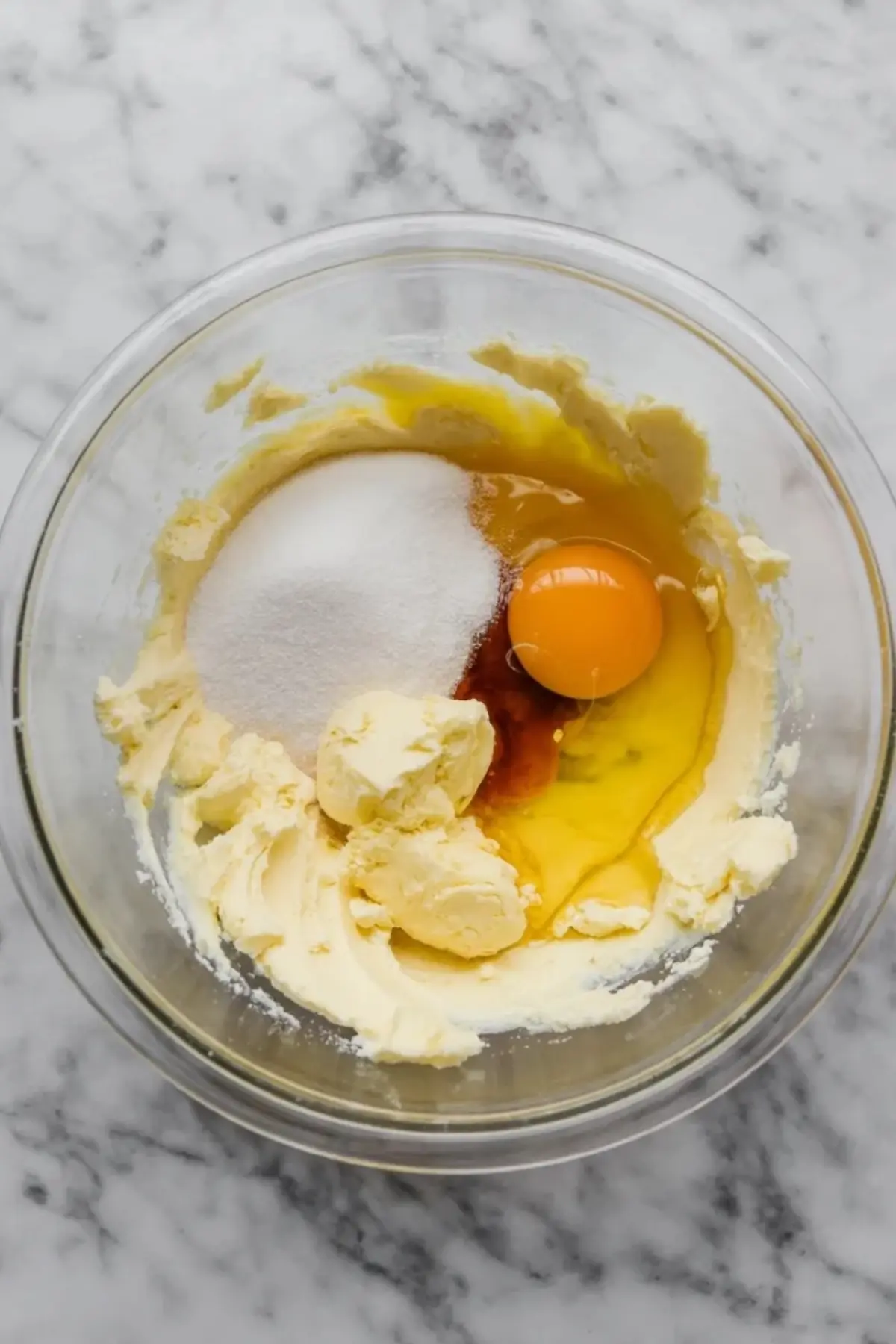 Butter, granulated sugar, a whole egg, and vanilla extract sit in a glass mixing bowl, showing the wet ingredients for a homemade sugar cookie recipe.
