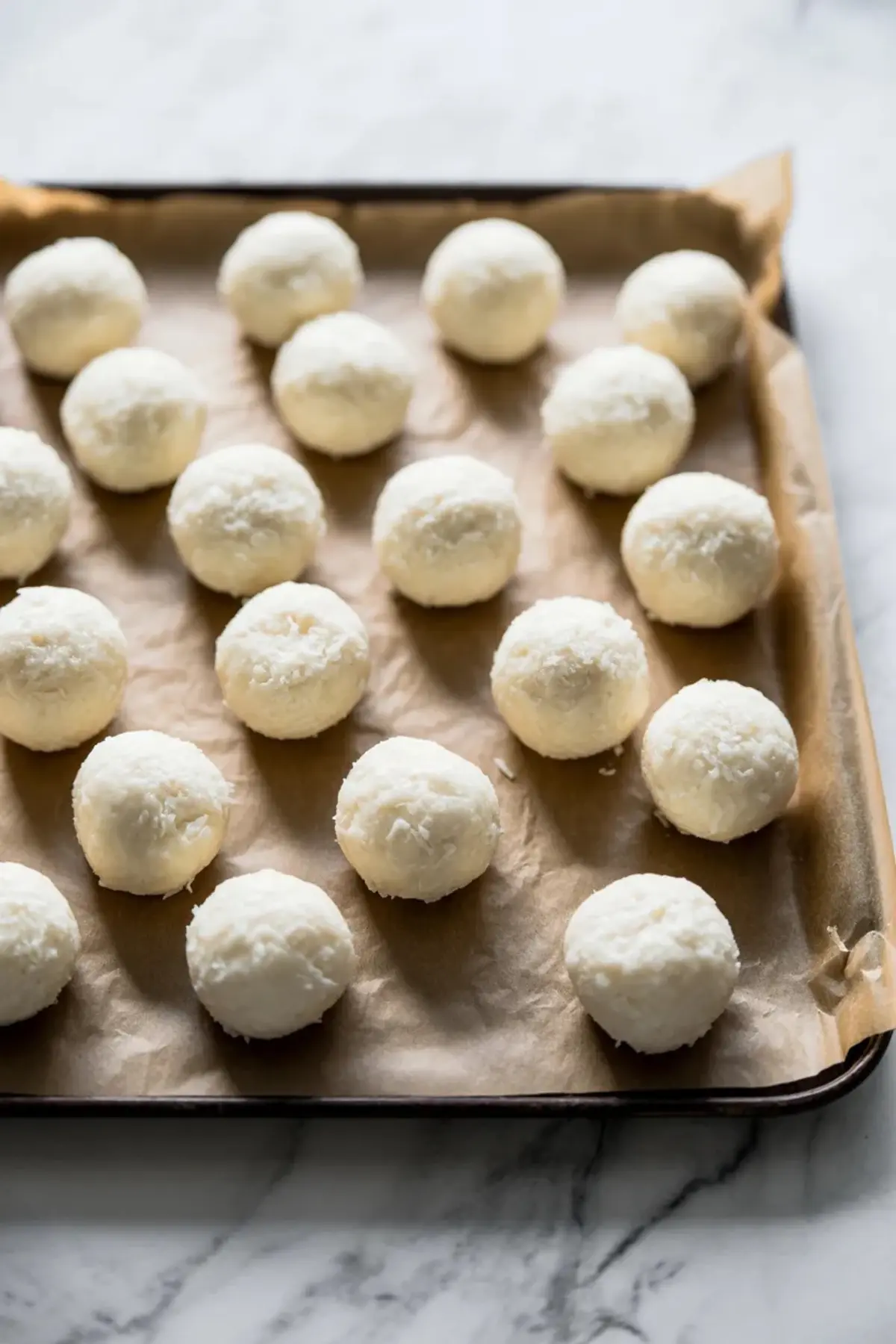 White coconut balls sit on a parchment lined baking sheet. The truffles have a smooth round shape and a shredded coconut coating. The tray rests on a marble countertop. The image shows a no bake coconut dessert recipe with easy homemade candy balls for Easter treats.
