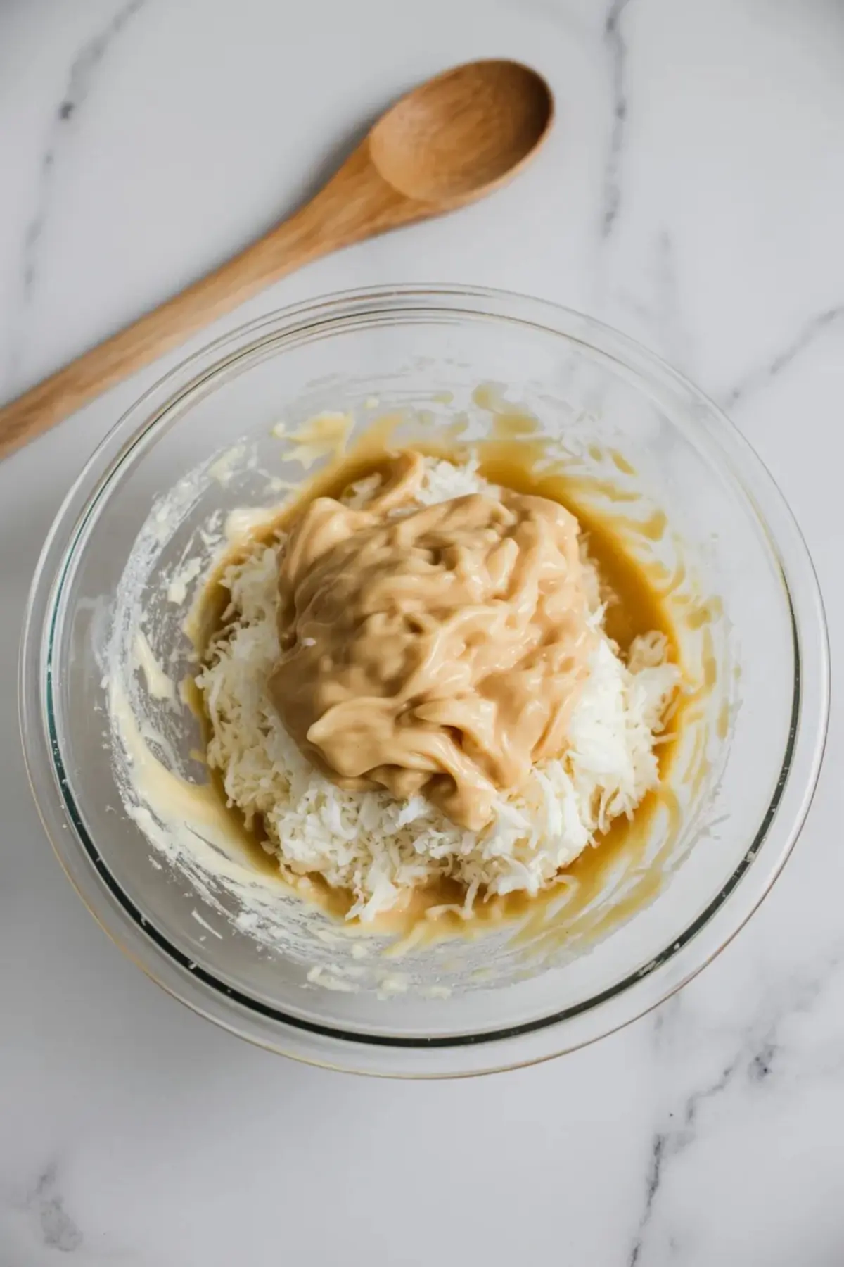 A glass mixing bowl holds shredded coconut and sweetened condensed milk. A wooden spoon rests beside the bowl. The mixture looks thick and creamy. The image shows simple ingredients for no bake coconut truffles and easy Easter dessert prep.
