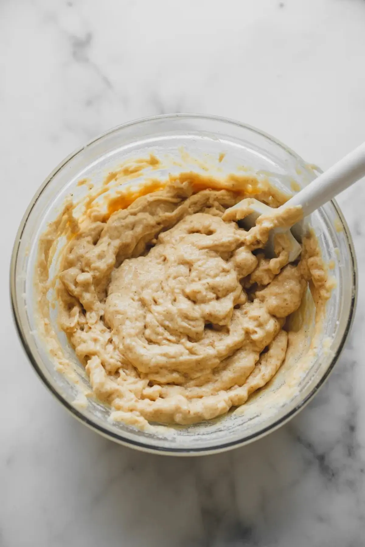 A glass bowl holds a thick coconut and sweetened condensed milk mixture. A spatula rests inside the bowl. The mixture looks creamy and sticky. The image shows step by step preparation for no bake coconut balls and easy Easter dessert recipe.

