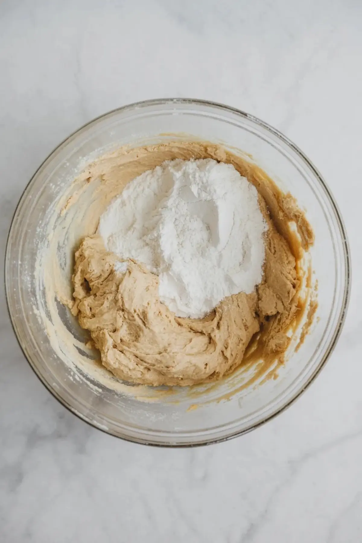 Glass mixing bowl holds thick sugar cookie dough with white flour added on top. The bowl sits on a white marble counter. The image shows Easter funfetti cookie dough in the mixing stage before stirring.
