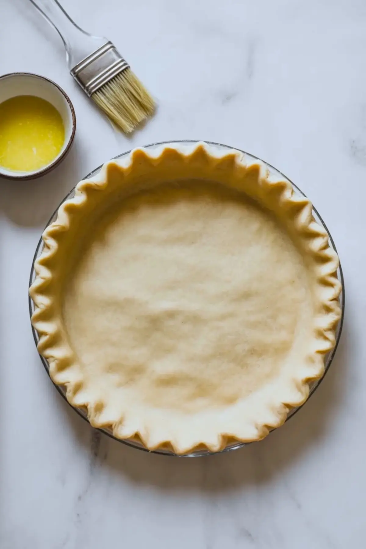 An unbaked pie crust sits in a glass pie dish with crimped edges on a white marble surface beside a small bowl of melted butter and a pastry brush, showing pie crust preparation for a homemade Easter swirl pie.