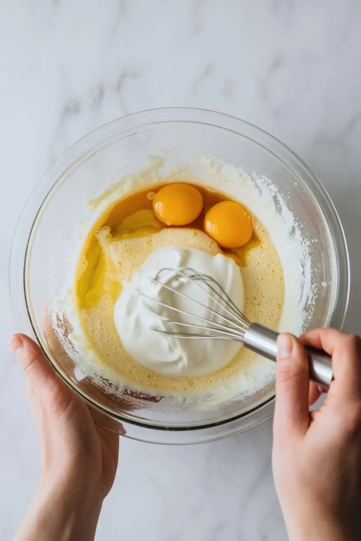 Hands whisk eggs and yogurt in a clear glass mixing bowl with melted butter for gluten free baked donut holes batter on a light marble countertop.
