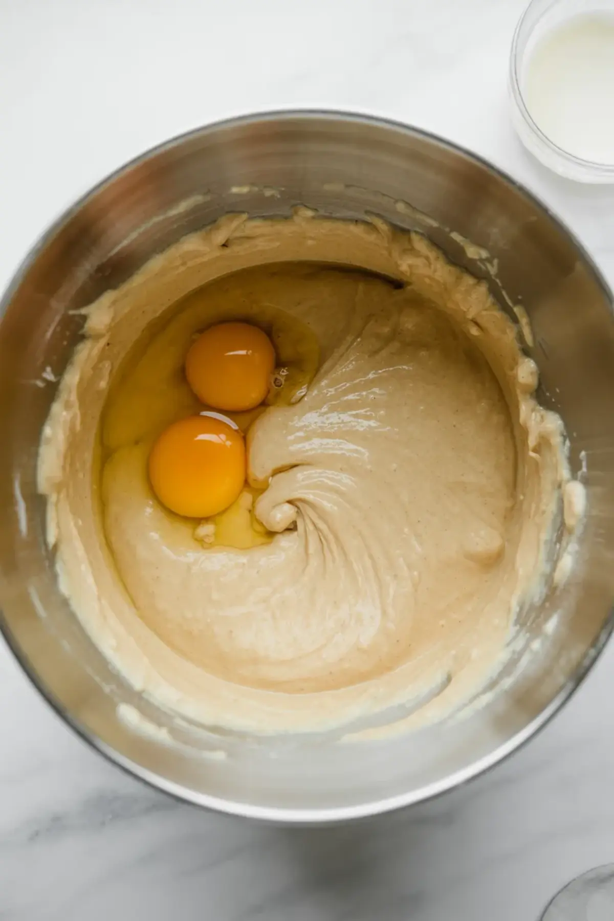 Two eggs sit in a smooth butter and sugar mixture inside a metal mixing bowl, ready to be blended into a rich cake batter. This step highlights gluten free coffee cake preparation and classic baking technique for moist cinnamon swirl cake.