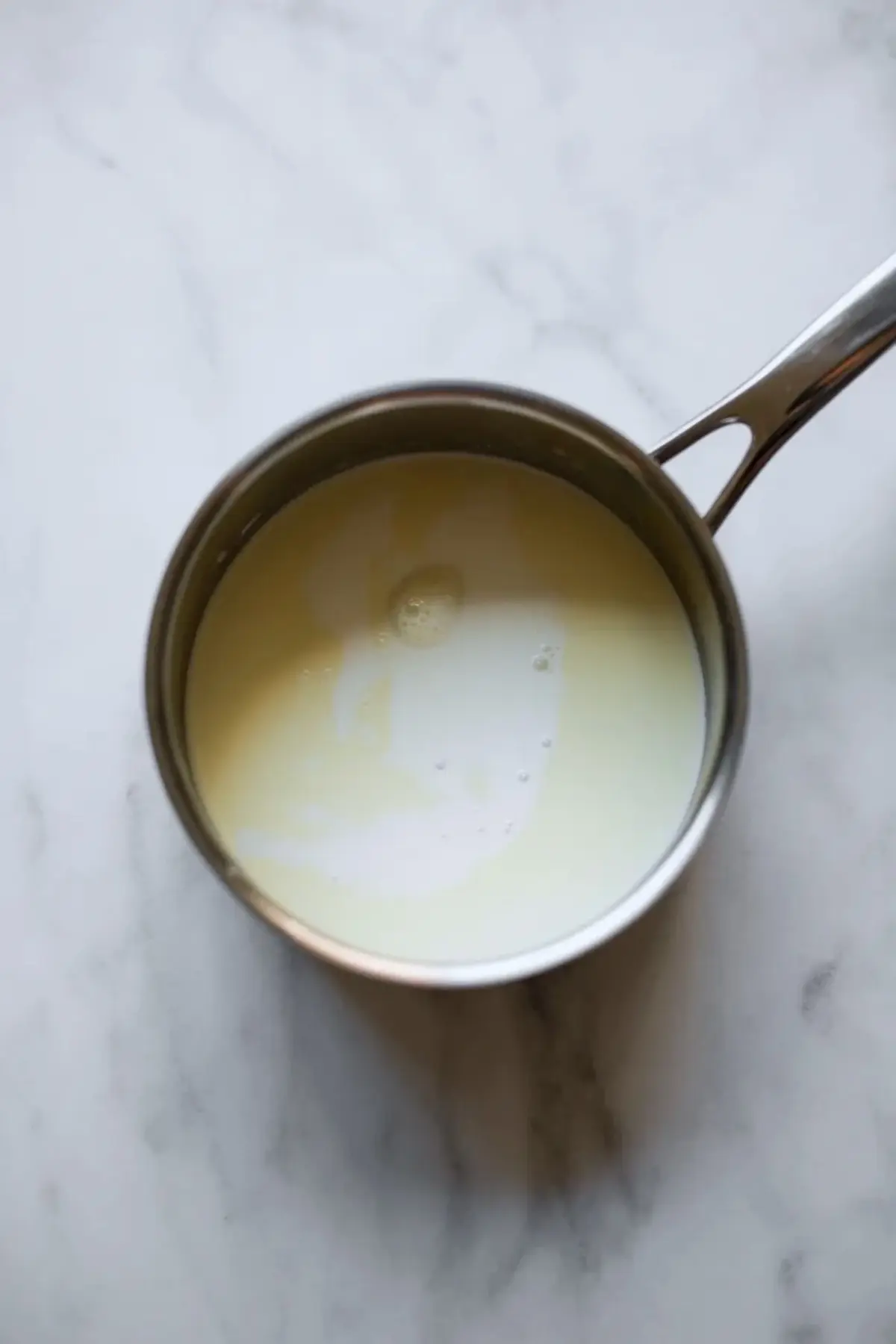 Milk and cream heat in a small stainless steel saucepan on a marble surface. The pale mixture shows light bubbles on top. This step-by-step image shows the base for homemade coffee creamer and an easy stovetop recipe.