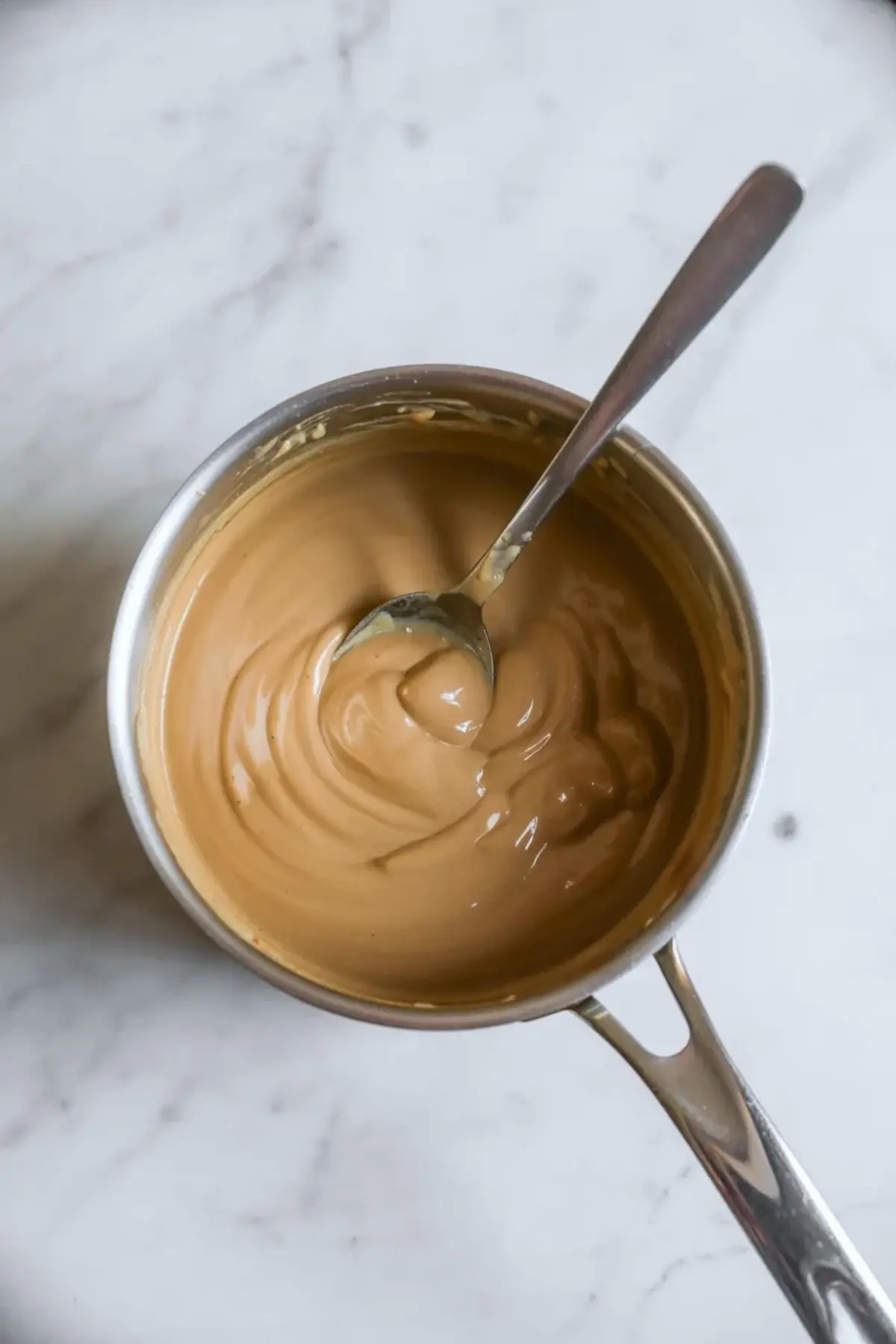 Thick caramel-colored coffee creamer fills a small saucepan with a spoon stirring the smooth mixture. The glossy texture shows a rich and creamy consistency. This image shows the cooking process for homemade vanilla coffee creamer.