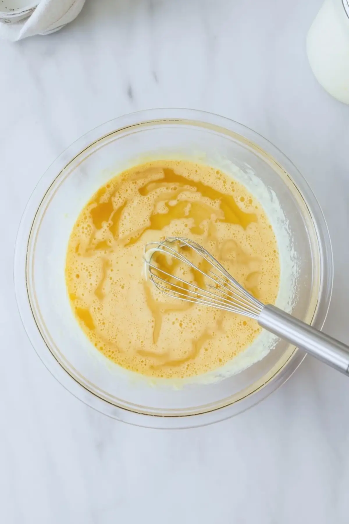 Glass bowl with whisked eggs and buttermilk mixture on a white marble surface, part of a homemade cornbread recipe preparation.

