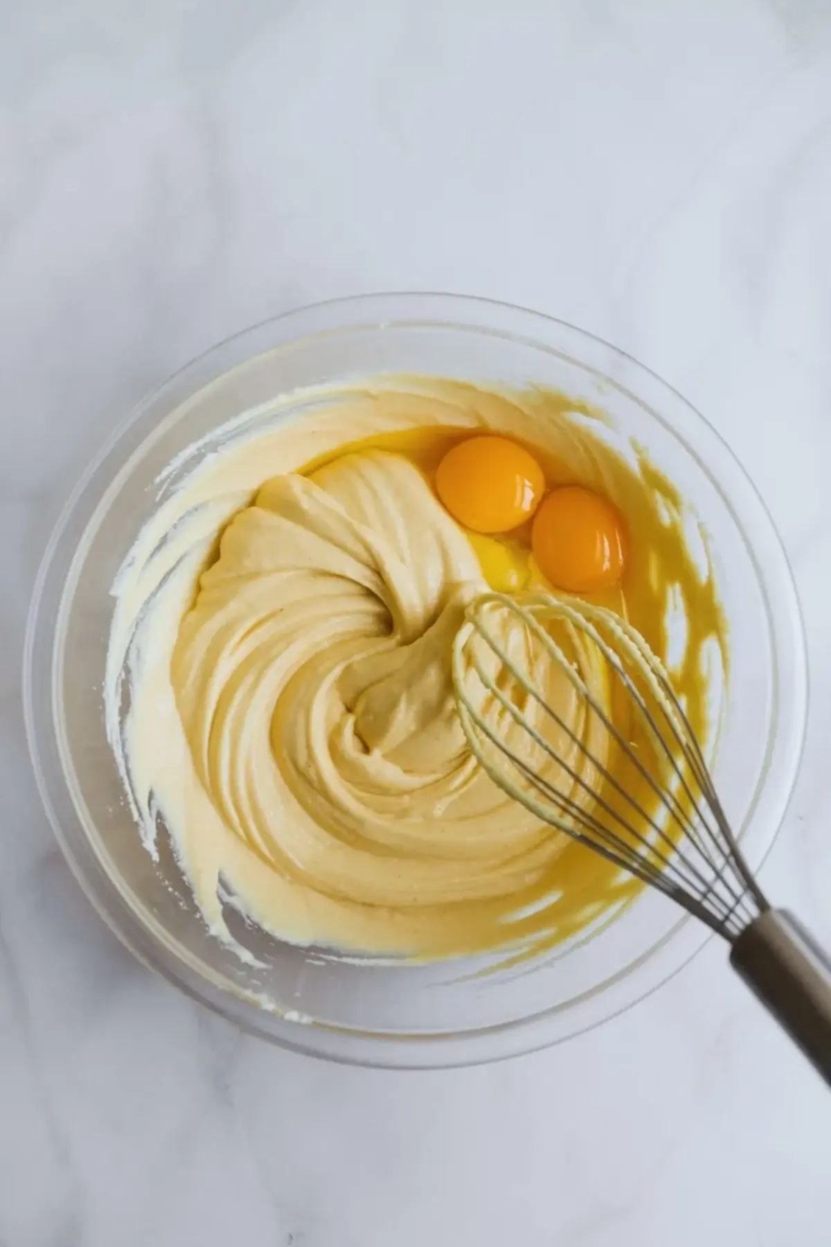 Glass mixing bowl with smooth cream cheese batter and two raw eggs on top. Metal whisk rests in the bowl. Thick pale yellow cheesecake mixture shows creamy texture during homemade Japanese cheesecake preparation on a marble surface.
