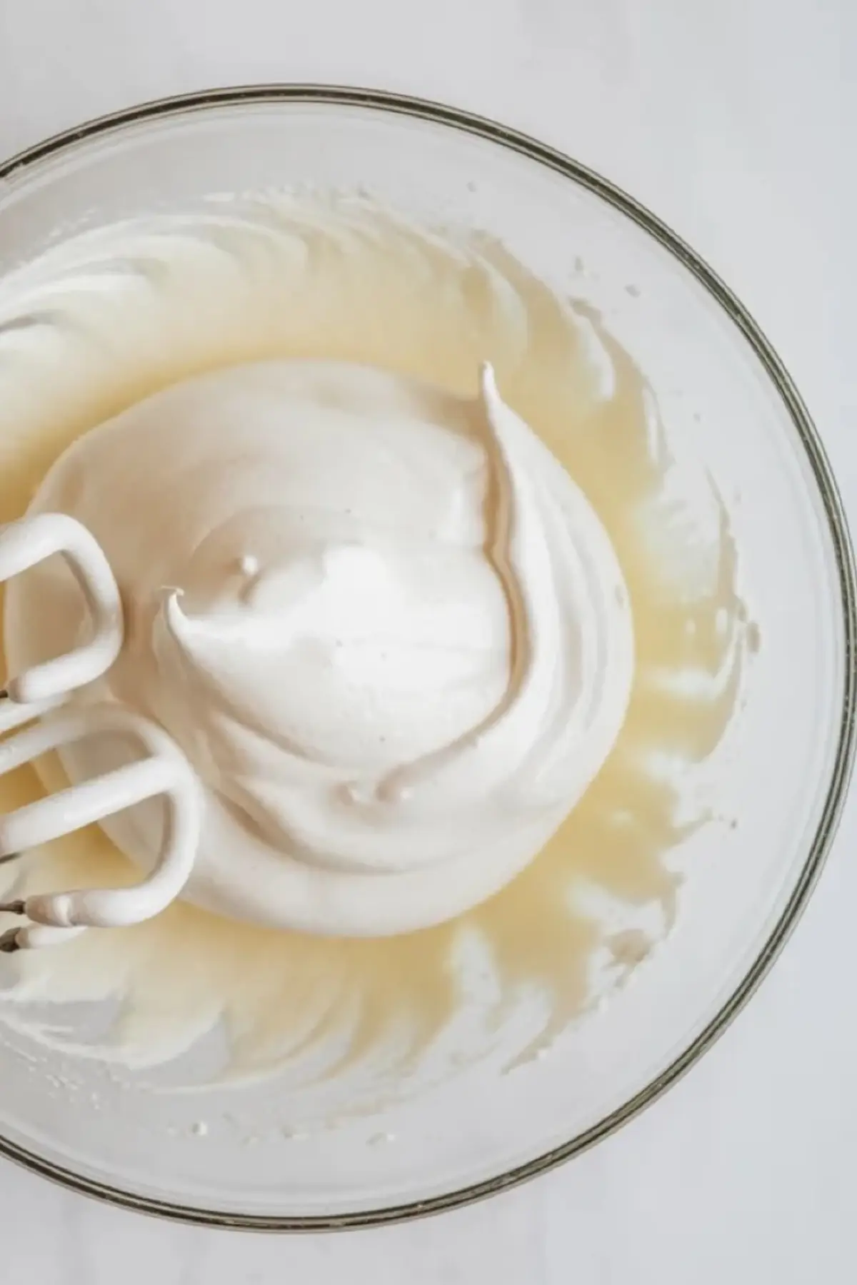 Glass bowl with glossy whipped egg whites forming soft peaks. Electric hand mixer beaters sit inside fluffy meringue. White airy foam shows volume and structure for Japanese cotton cheesecake recipe preparation.