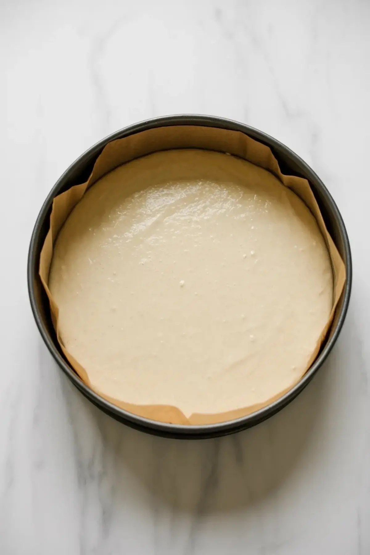 Japanese cheesecake batter in a round springform pan lined with parchment paper. Smooth pale cream batter fills the pan evenly. Overhead view shows raw cheesecake mixture before baking on a white marble surface.