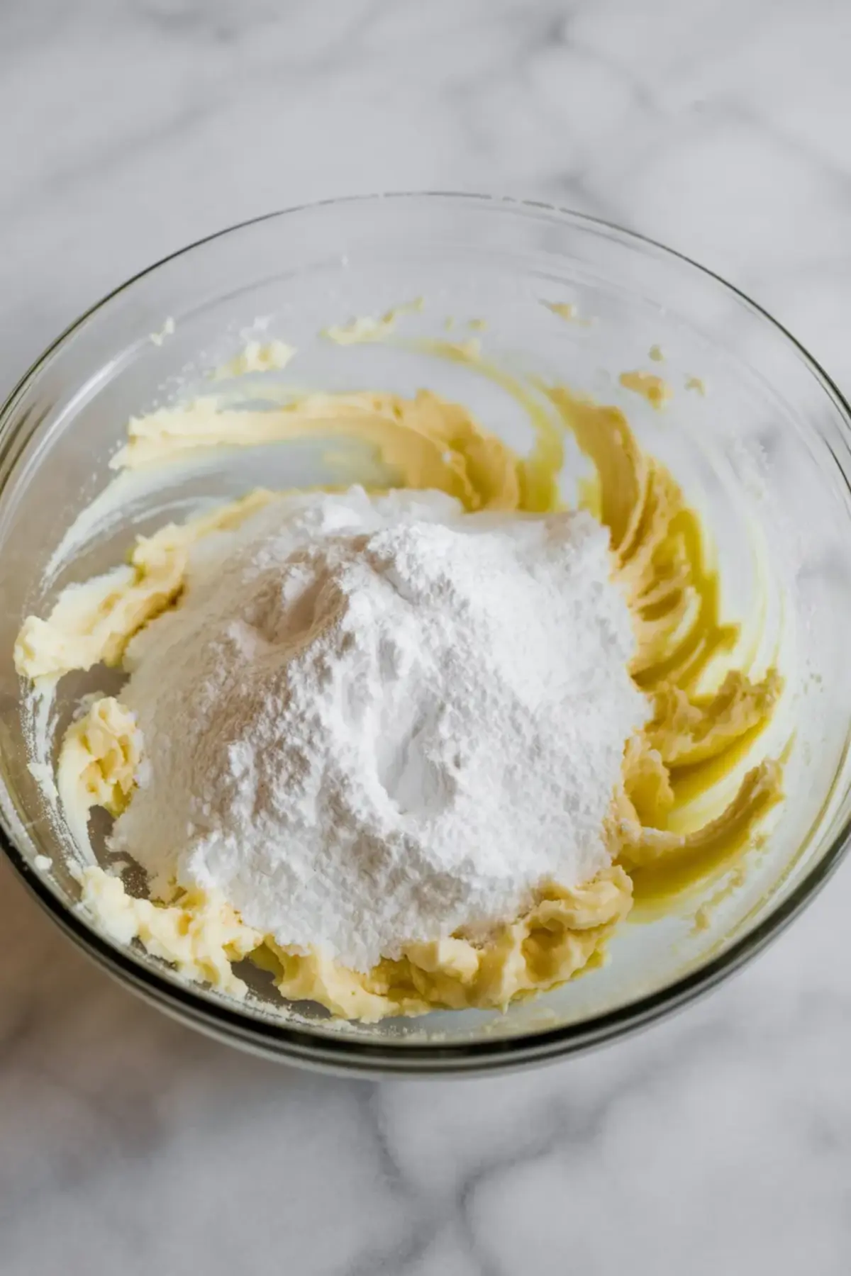 Glass mixing bowl holds creamed butter topped with powdered sugar on a marble countertop. Thick yellow butter mixture surrounds a mound of white sugar, showing the start of homemade espresso buttercream frosting.