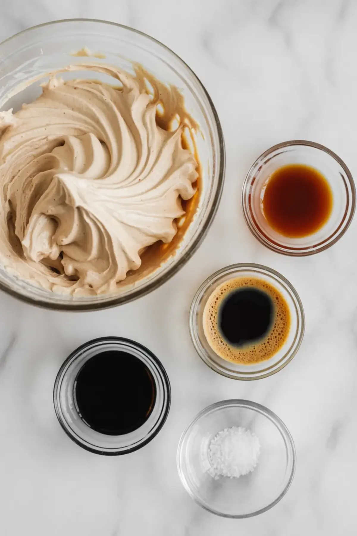 Top view shows a bowl of whipped espresso buttercream surrounded by small glass bowls of brewed espresso, coffee liqueur, and salt on a marble surface. Ingredients display supports a homemade Kahlua espresso buttercream recipe.