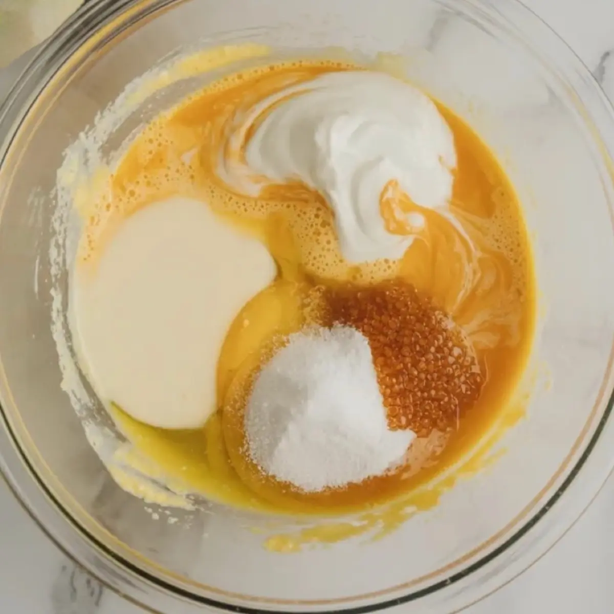 Wet ingredients for keto cornbread in a glass mixing bowl, eggs, sour cream, sweetener, and melted butter partially mixed into a yellow batter.
