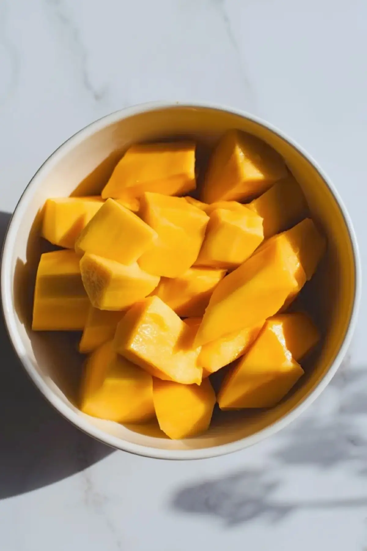 Fresh mango chunks fill a white bowl on a light marble surface. Bright yellow mango pieces show smooth texture and clean cuts, creating a vibrant tropical fruit preparation scene for homemade mango ice cream.
