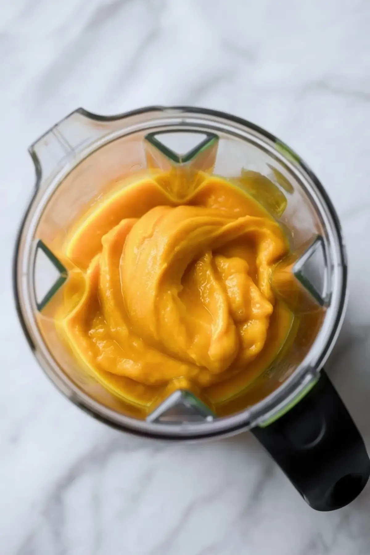 Blender jar holds smooth mango puree with a thick and creamy texture. Bright orange mango mixture swirls inside the blender, showing a fresh fruit base for easy mango ice cream.
