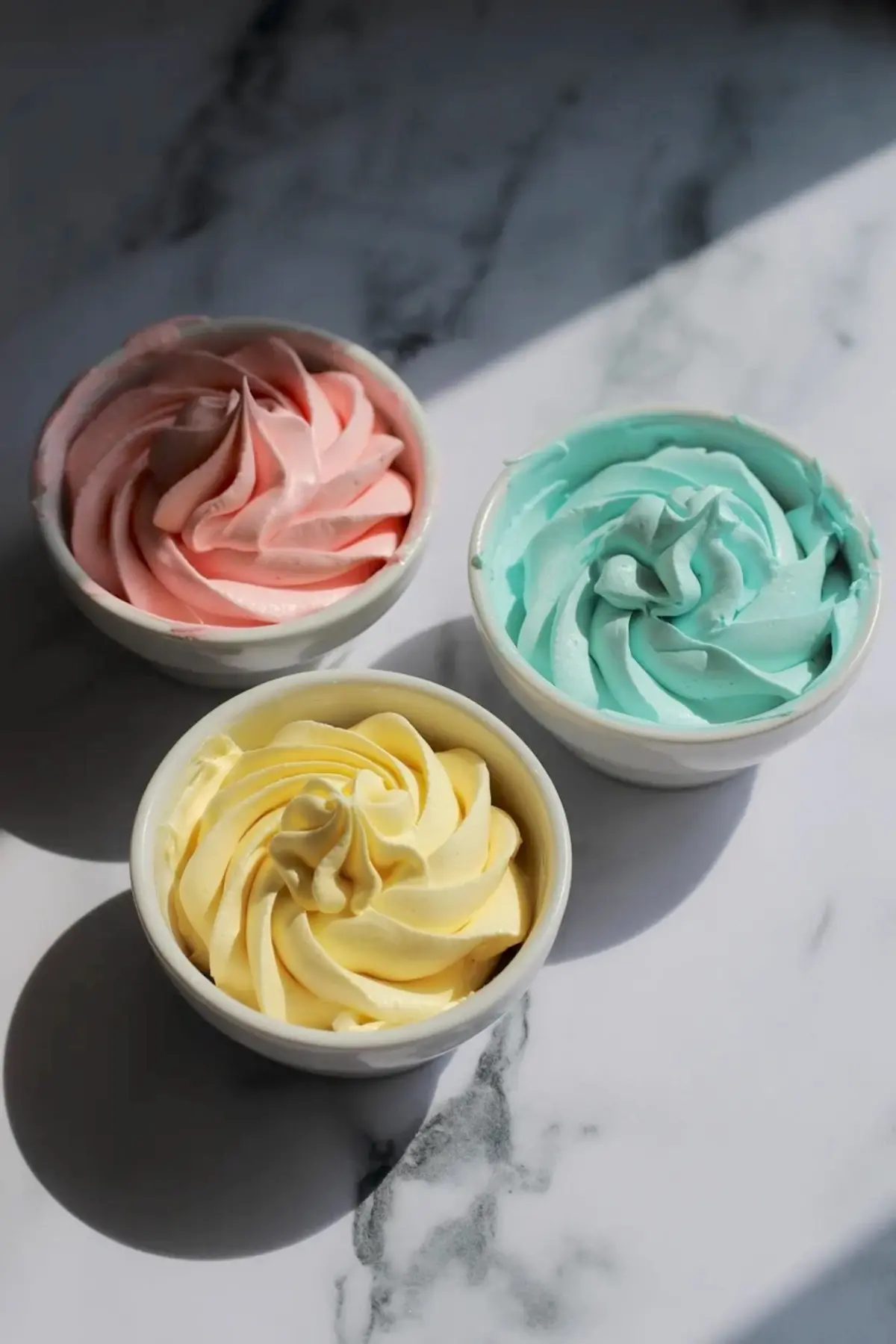 Three small white bowls with piped pastel buttercream frosting in pink, blue, and yellow shades, arranged on a sunlit marble surface.
