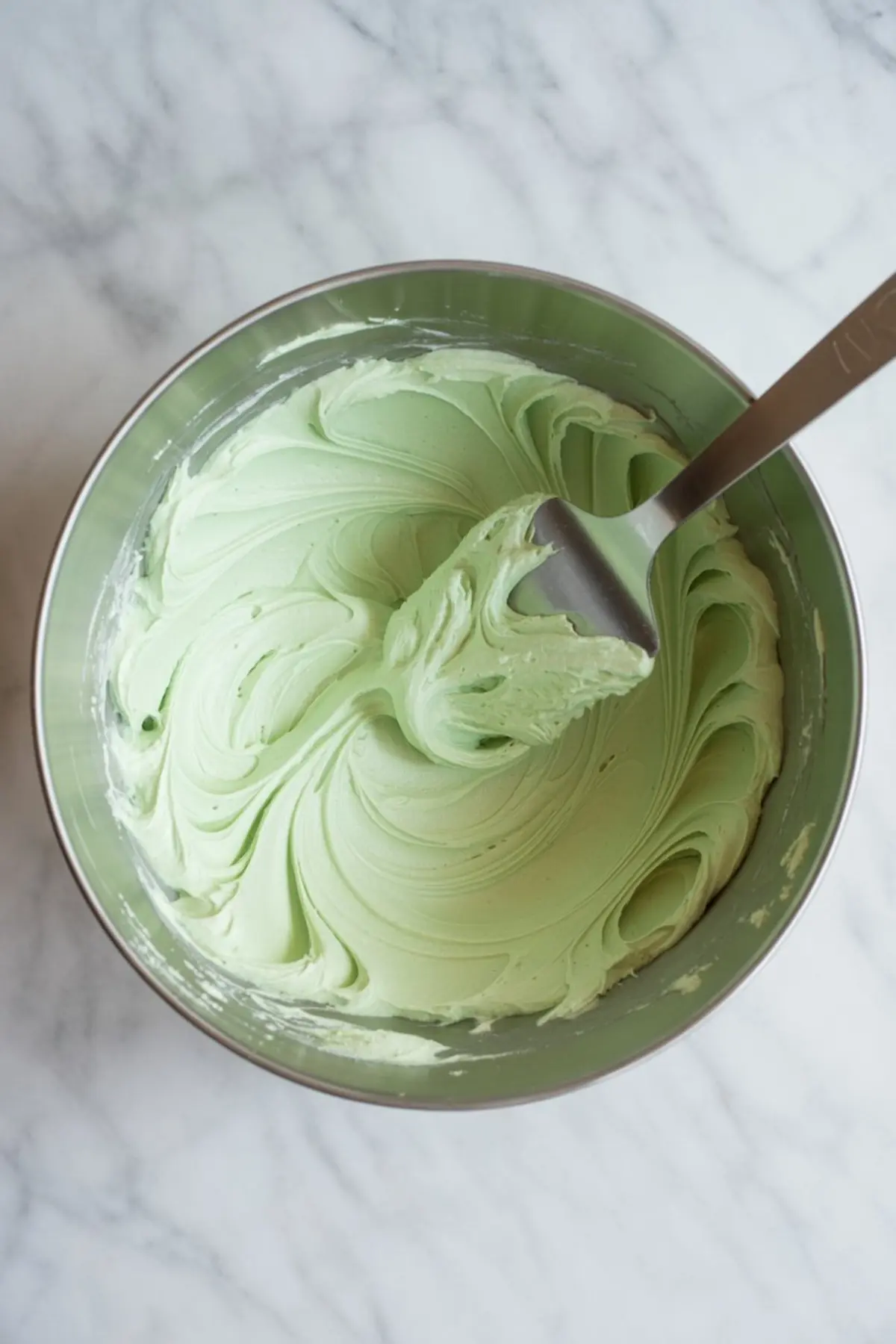 A metal mixing bowl holds pale green mint buttercream frosting. A spatula lifts the smooth frosting to show a creamy texture. The bowl sits on a white marble surface. This image shows homemade mint frosting for mint chocolate chip cake.