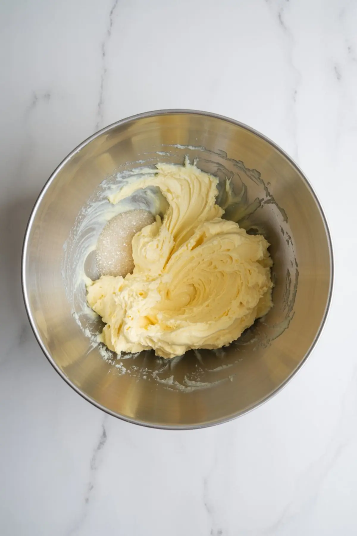 A metal mixing bowl holds creamed butter and sugar with visible sugar granules. The mixture looks pale yellow and fluffy. The bowl sits on a white marble surface. This image shows the butter and sugar creaming step in cake baking.