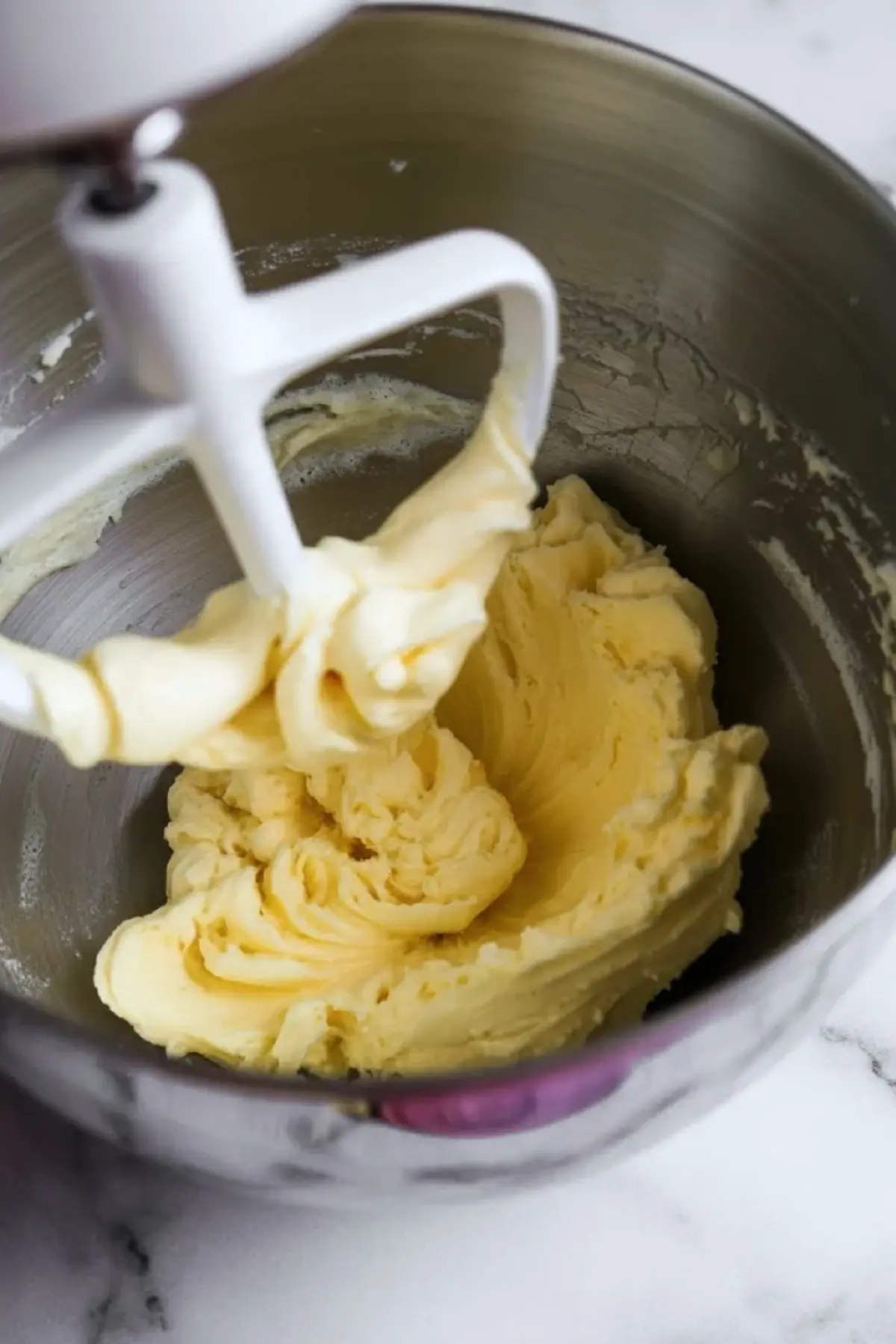 Creamed butter blends inside a stainless steel stand mixer bowl with a paddle attachment, showing smooth yellow butter texture for cake batter preparation.
