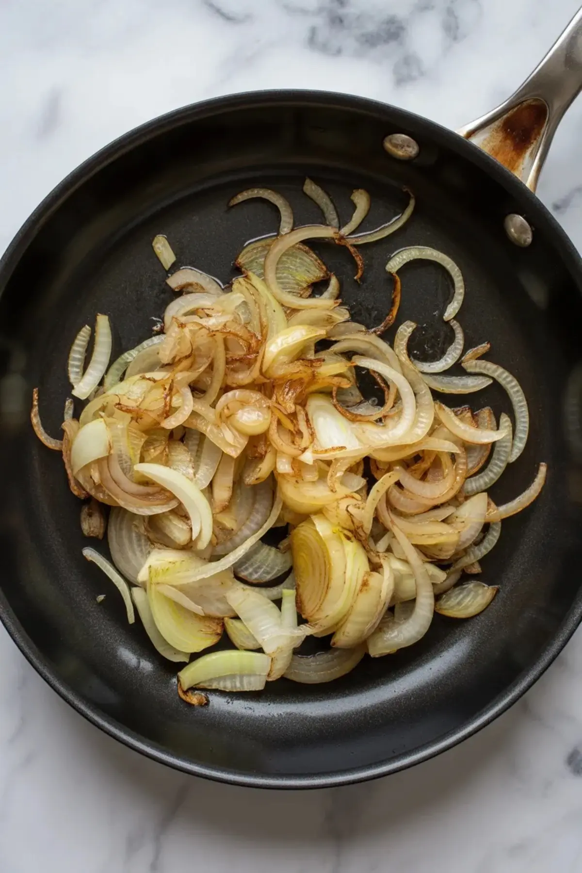 Sliced onions slowly caramelizing in a black skillet on a marble countertop, showing a golden-brown sear ideal for making savory onion jam.
