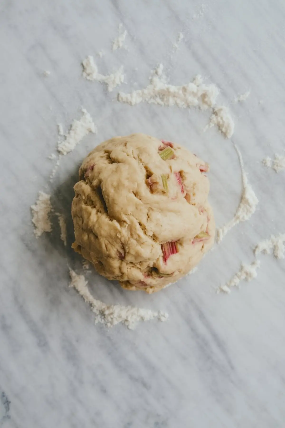 Rhubarb scone dough ball rests on a marble surface dusted with flour. Pink and green rhubarb pieces are visible throughout the soft, slightly sticky dough, showing a homemade pastry preparation process.