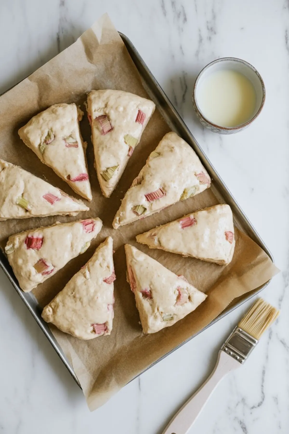 Unbaked triangular rhubarb scones sit on a parchment-lined baking sheet. A pastry brush and a small bowl of milk rest nearby, showing the egg wash step before baking.