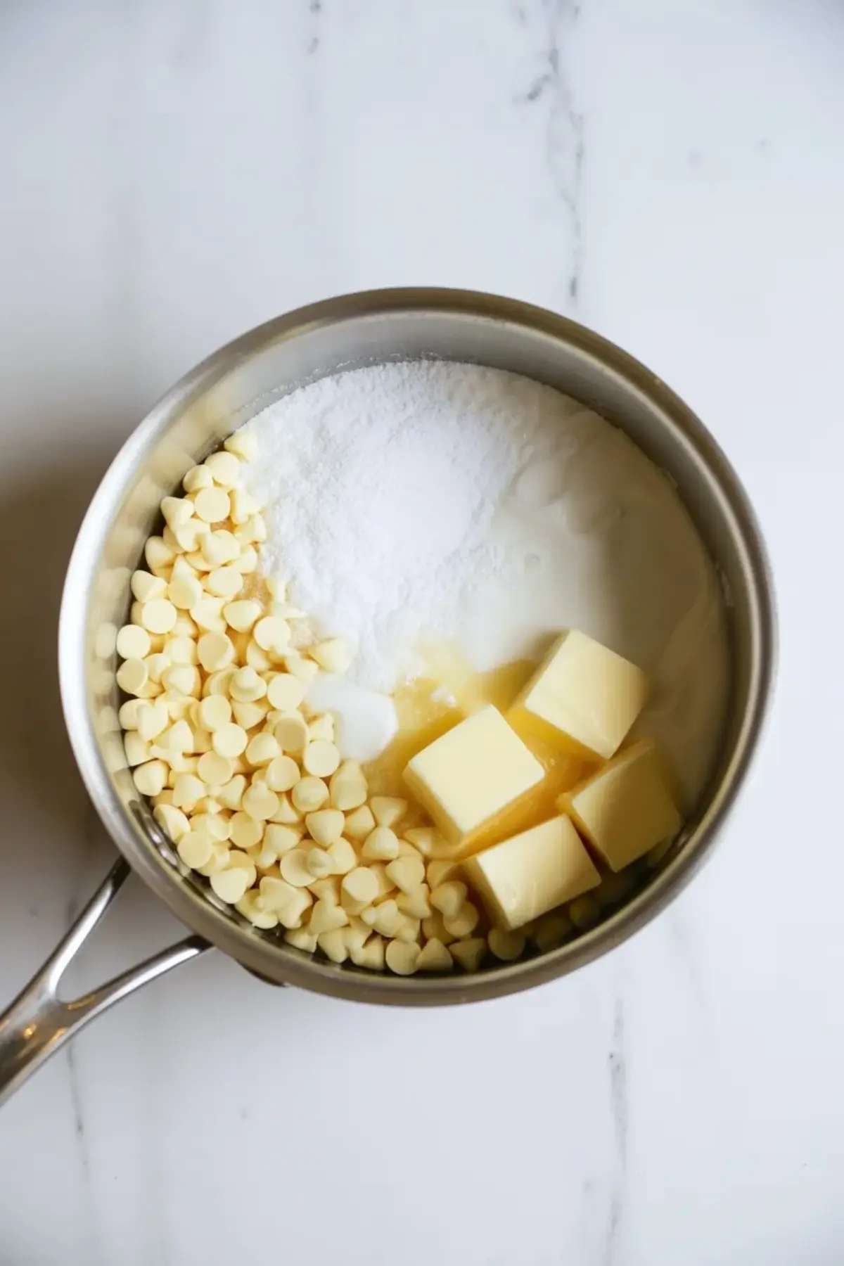 Saucepan holds white chocolate chips, granulated sugar, butter cubes, and sweetened condensed milk on a white marble countertop. Ingredients sit unmelted in a metal pot ready for stovetop fudge preparation.