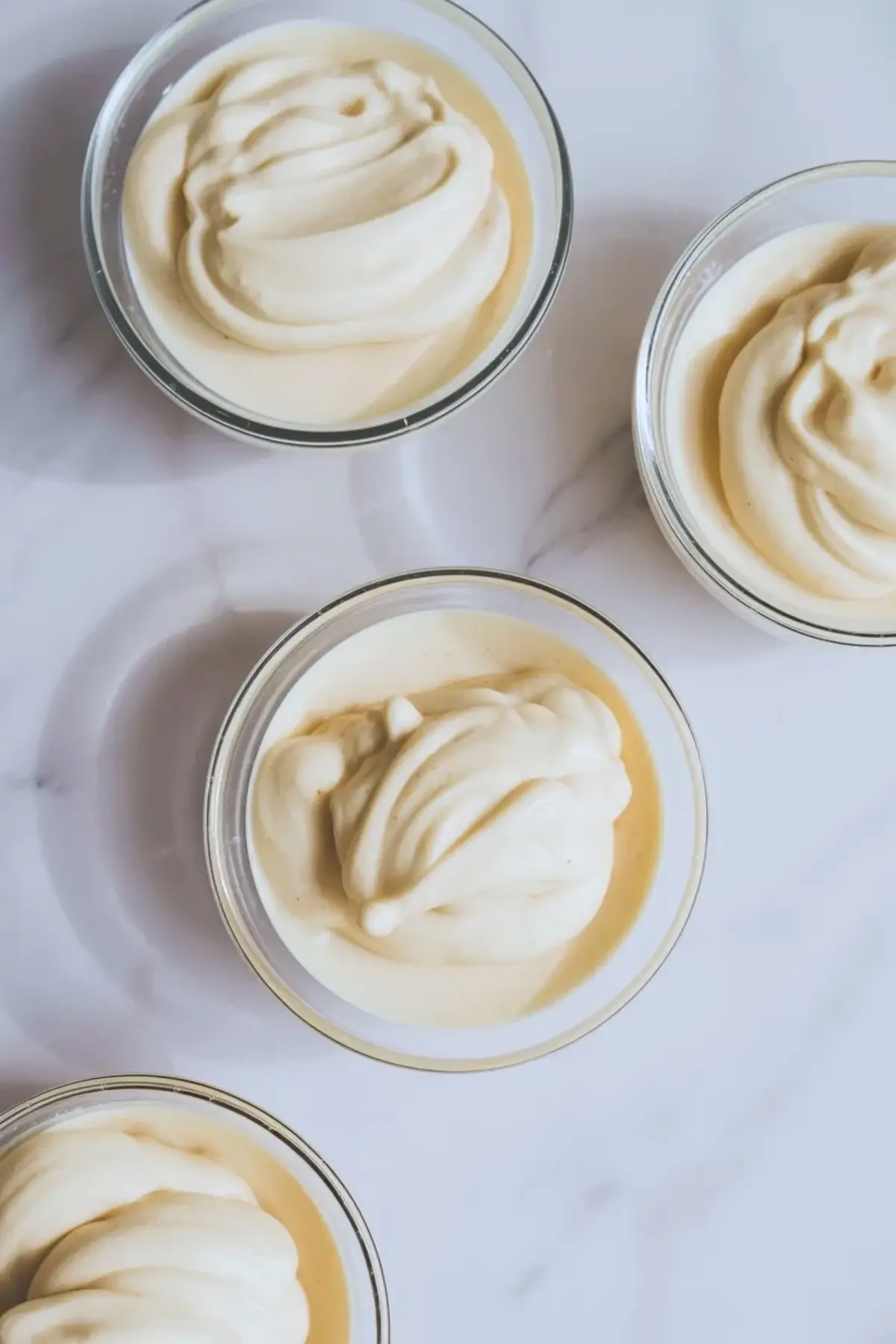 Small glass jars hold smooth vanilla fudge filling with a creamy swirled top. Jars sit spaced on a white marble background ready for layering or decorating.
