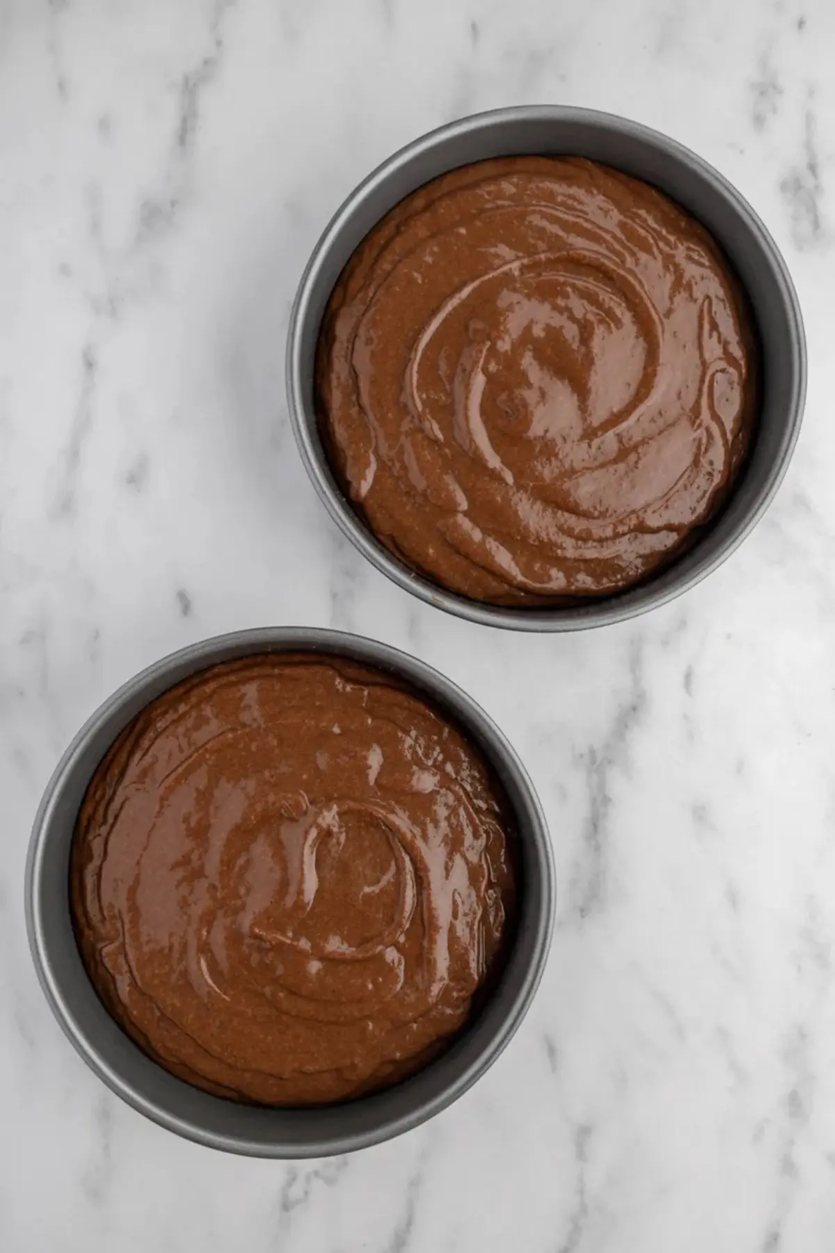 Two round cake pans filled with freshly mixed chocolate cake batter, placed on a marble surface and ready for baking, showcasing a smooth swirl texture.
