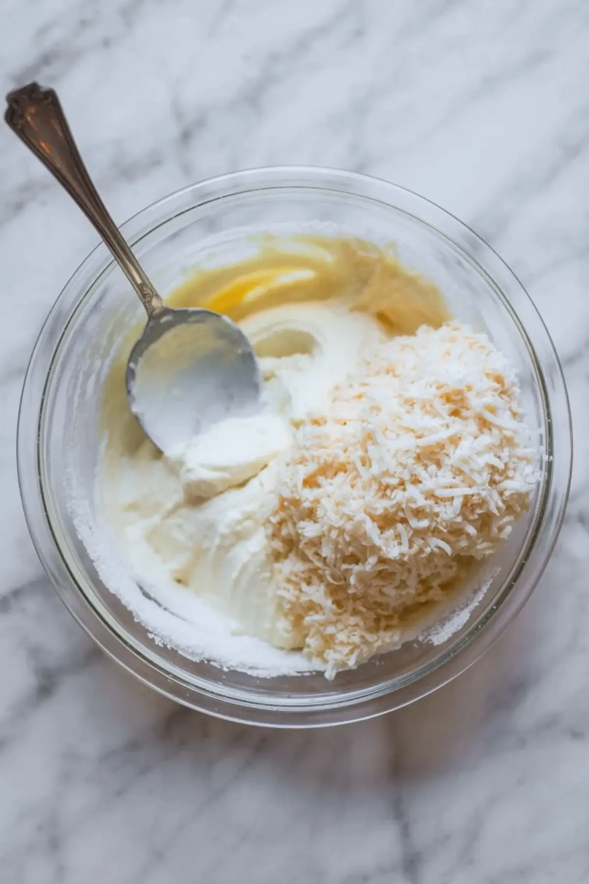 Glass mixing bowl holds sour cream, shredded coconut, sugar, and egg mixture. A spoon rests in the bowl, showing ingredients for sour cream coconut cake batter on a marble surface.
