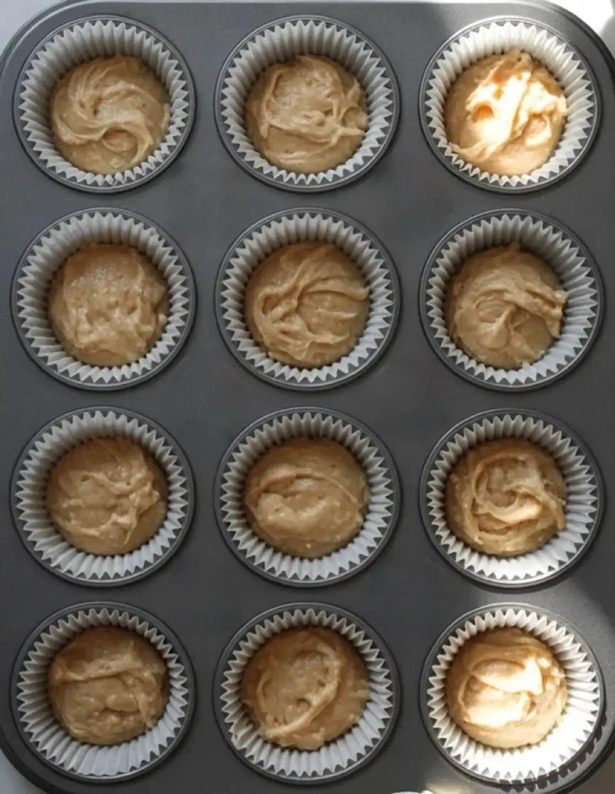 Muffin tin filled with unbaked sour cream muffin batter in paper liners, evenly portioned for baking, viewed from above on a neutral surface.
