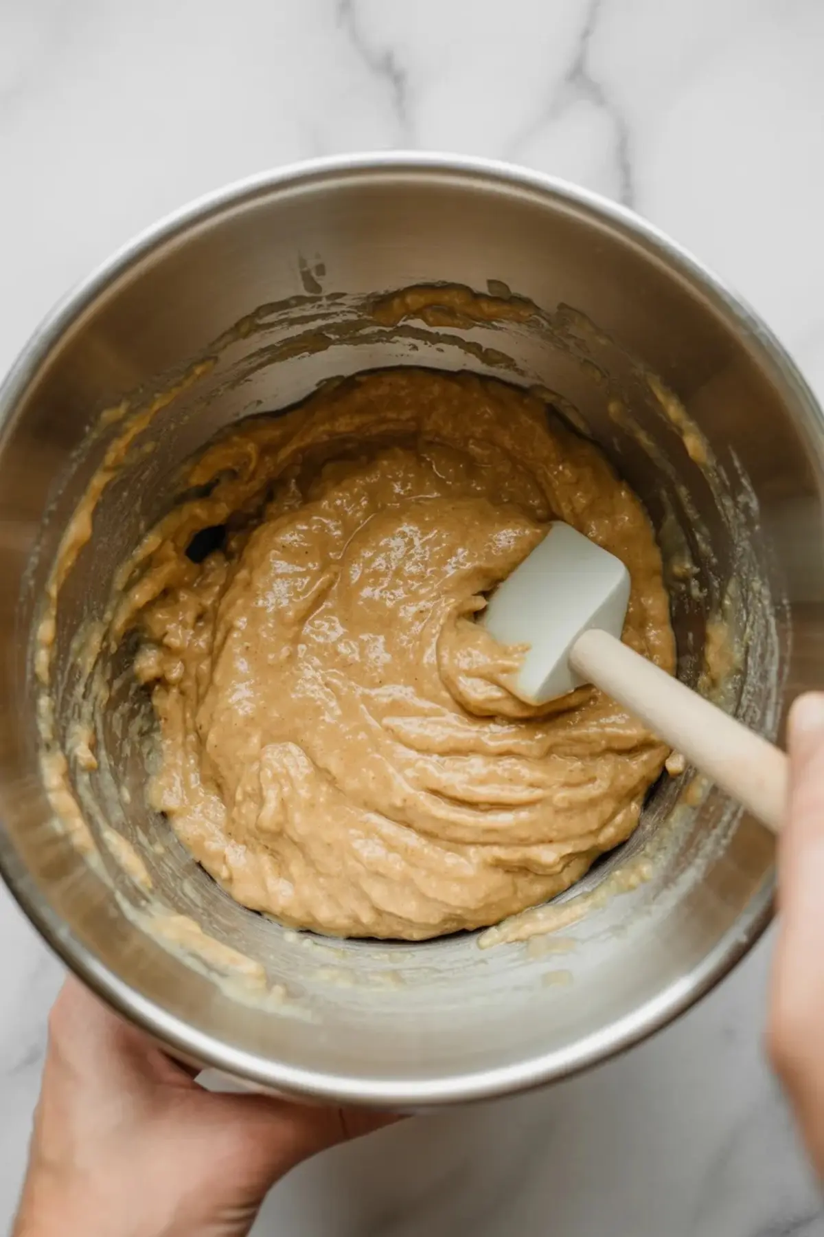 Hand holding a metal bowl of thick, well-mixed muffin batter with a white silicone spatula resting inside, set against a white marble background.

