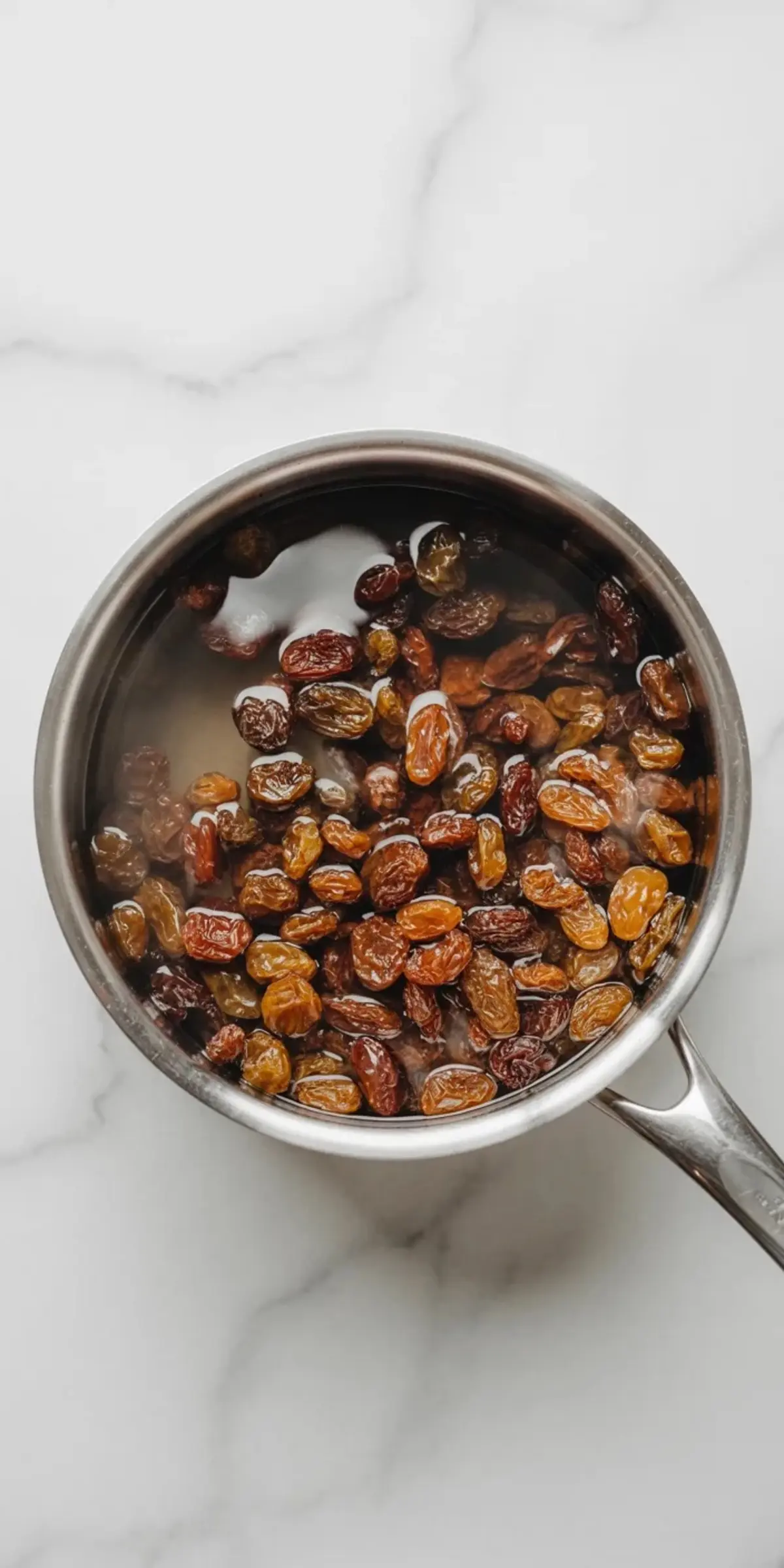 Metal saucepan holds raisins soaking in hot water on a white marble surface. Plump golden and dark raisins float in clear liquid, showing a simple step for softening dried fruit for baking and sour cream raisin pie filling.
