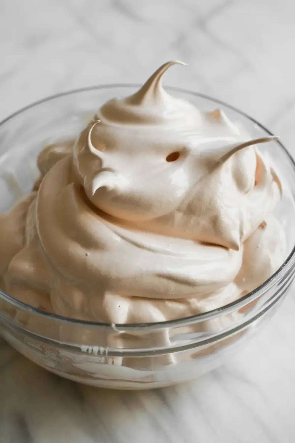 Glass bowl holds thick whipped sour cream mixture with soft peaks on a marble countertop. Creamy vanilla filling shows smooth texture for homemade sour cream raisin pie and classic custard dessert recipes.
