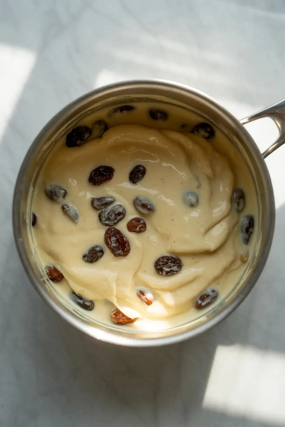 Metal saucepan contains creamy custard mixed with soaked raisins on a light marble surface. Glossy sour cream filling surrounds plump raisins, showing cooked pie filling for old fashioned sour cream raisin pie.
