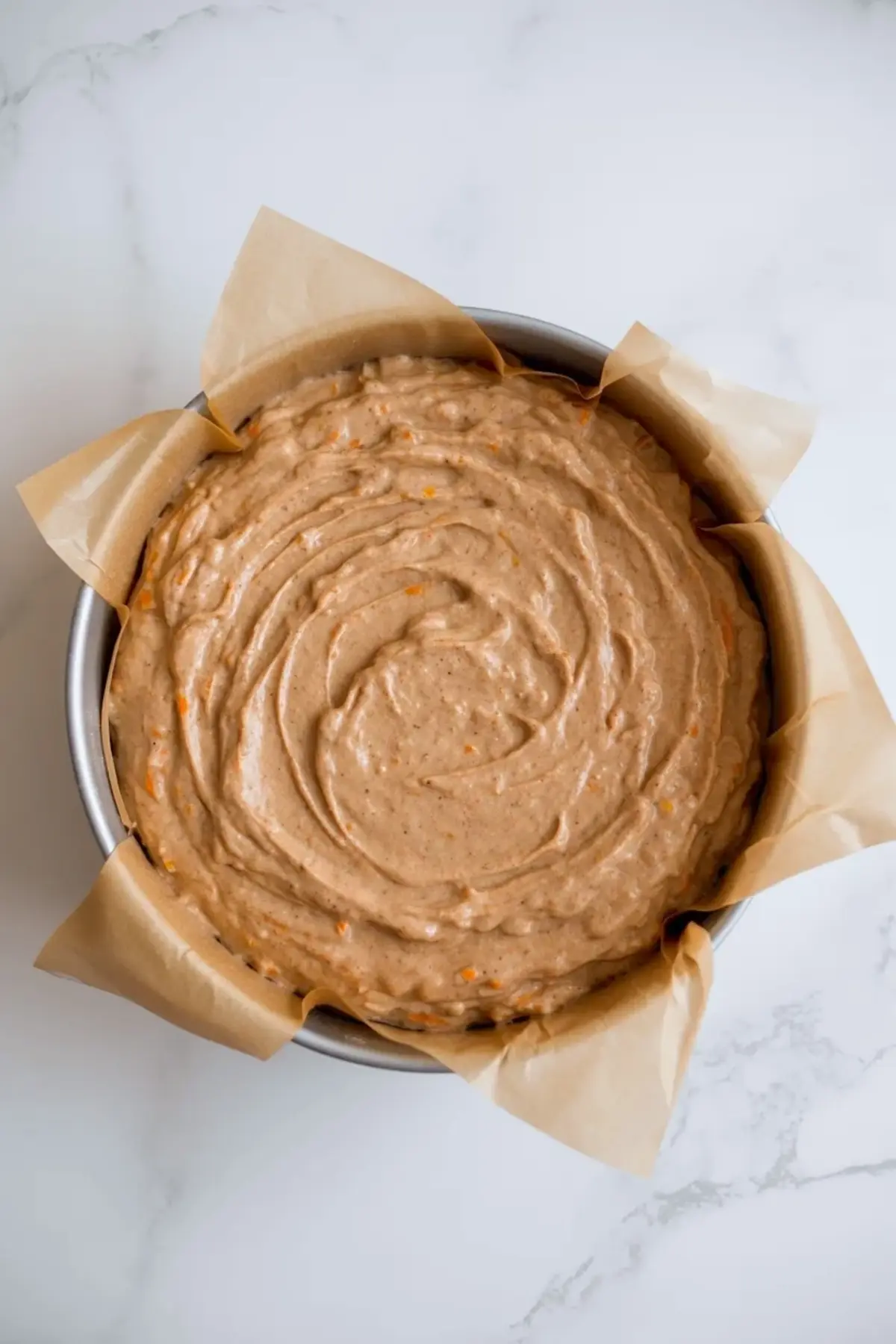 Overhead view of sourdough carrot cake batter swirled in a round cake pan lined with parchment paper, thick spiced batter with visible grated carrots on a white marble surface, step by step homemade cake recipe.