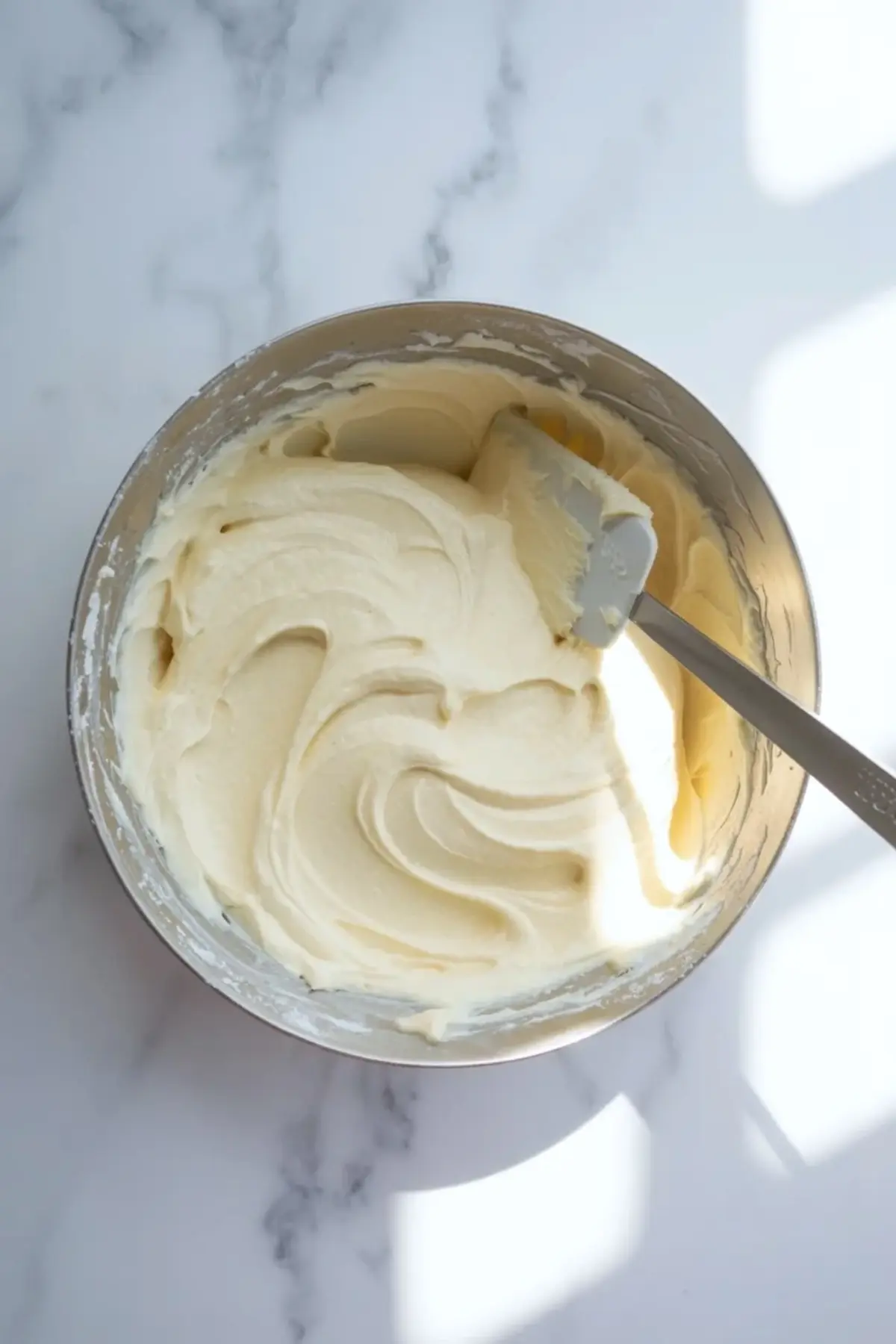 Smooth cream cheese frosting whipped in a glass bowl with a silicone spatula, thick and creamy vanilla frosting for homemade carrot cake on a bright marble background.