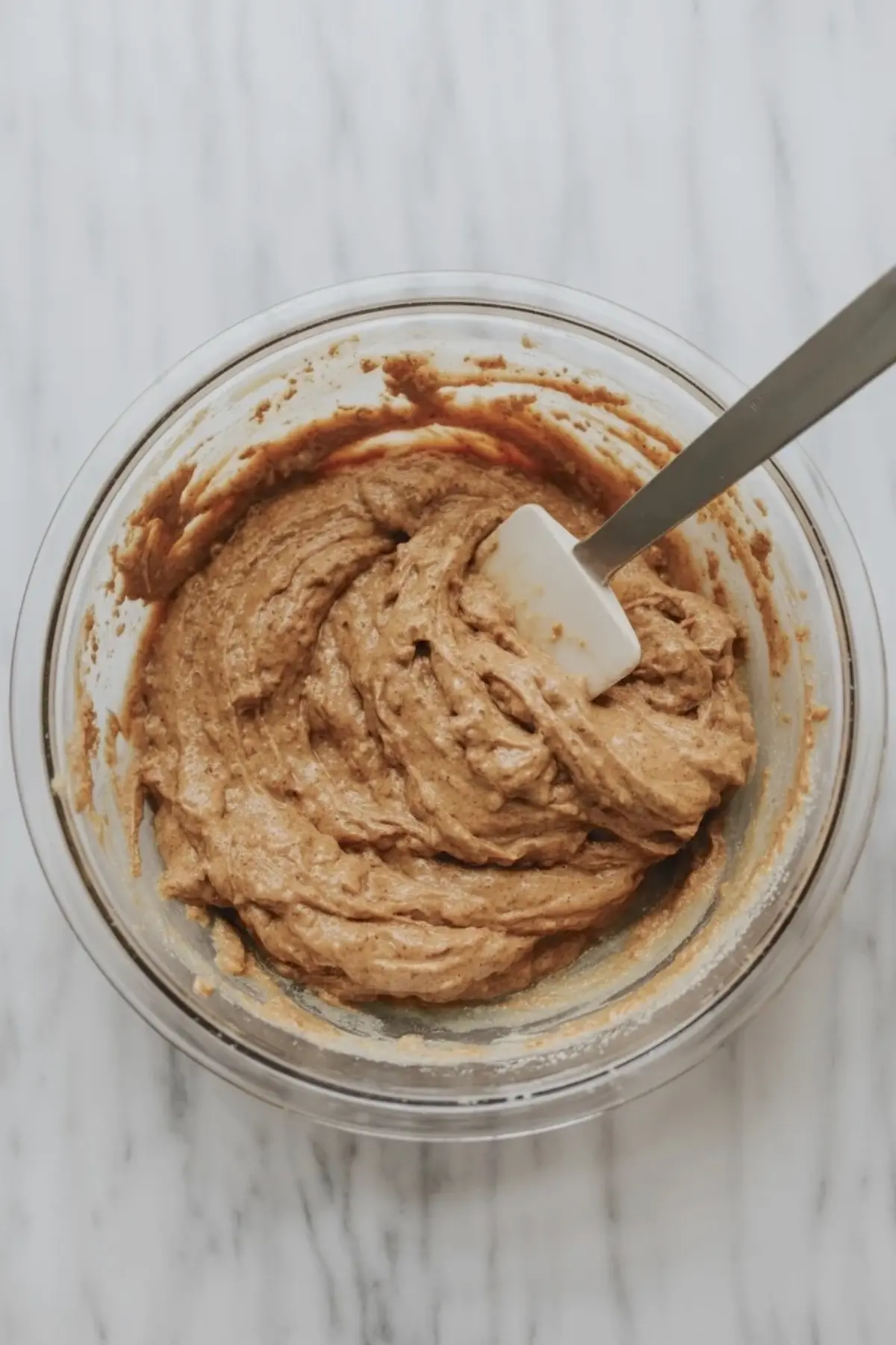 Thick sourdough carrot cake batter mixed in a glass bowl with a white spatula, rich brown spiced batter ready for baking on a marble countertop.