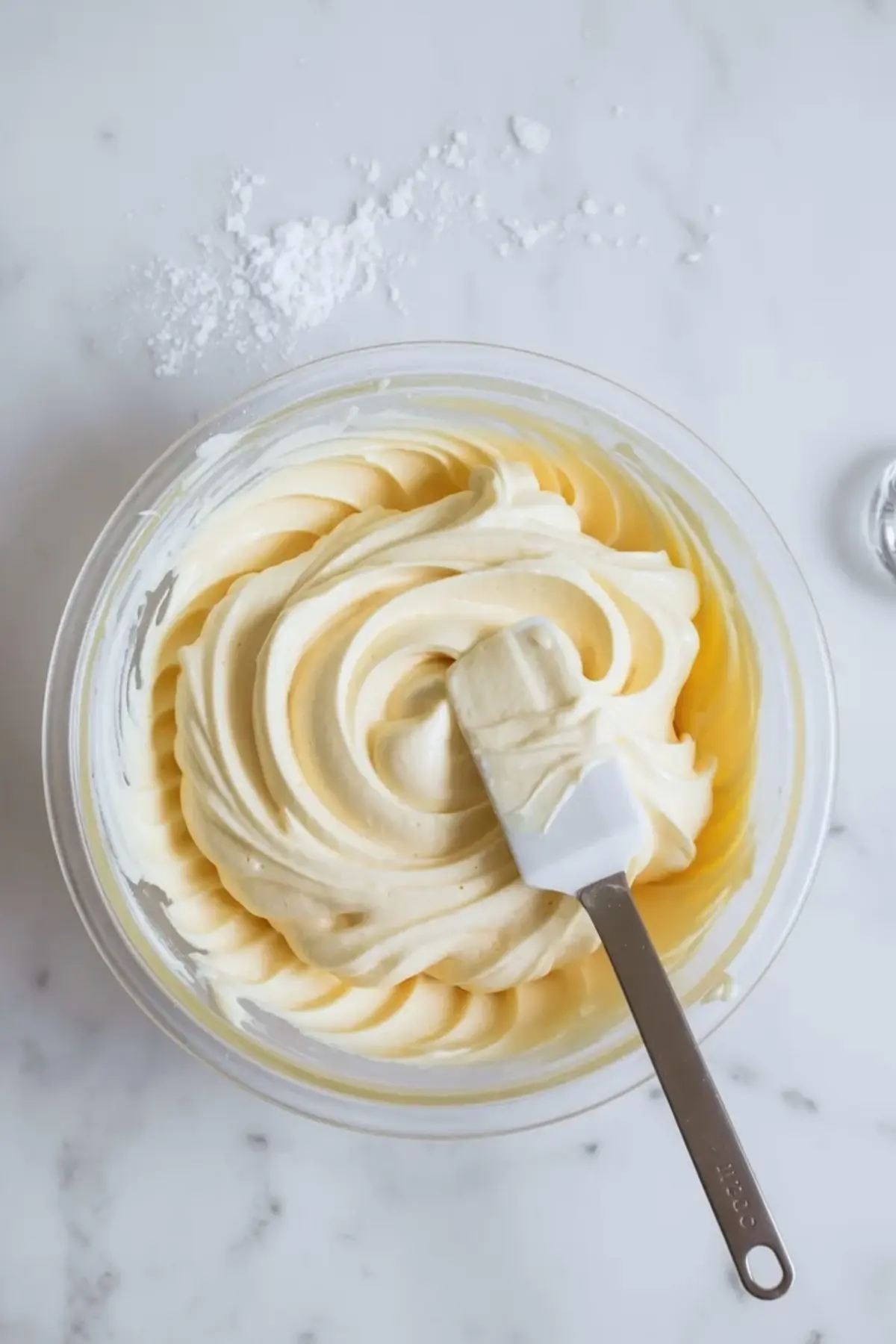 Creamy vanilla frosting swirled in a clear glass mixing bowl with a rubber spatula, resting on a marble surface dusted with powdered sugar.
