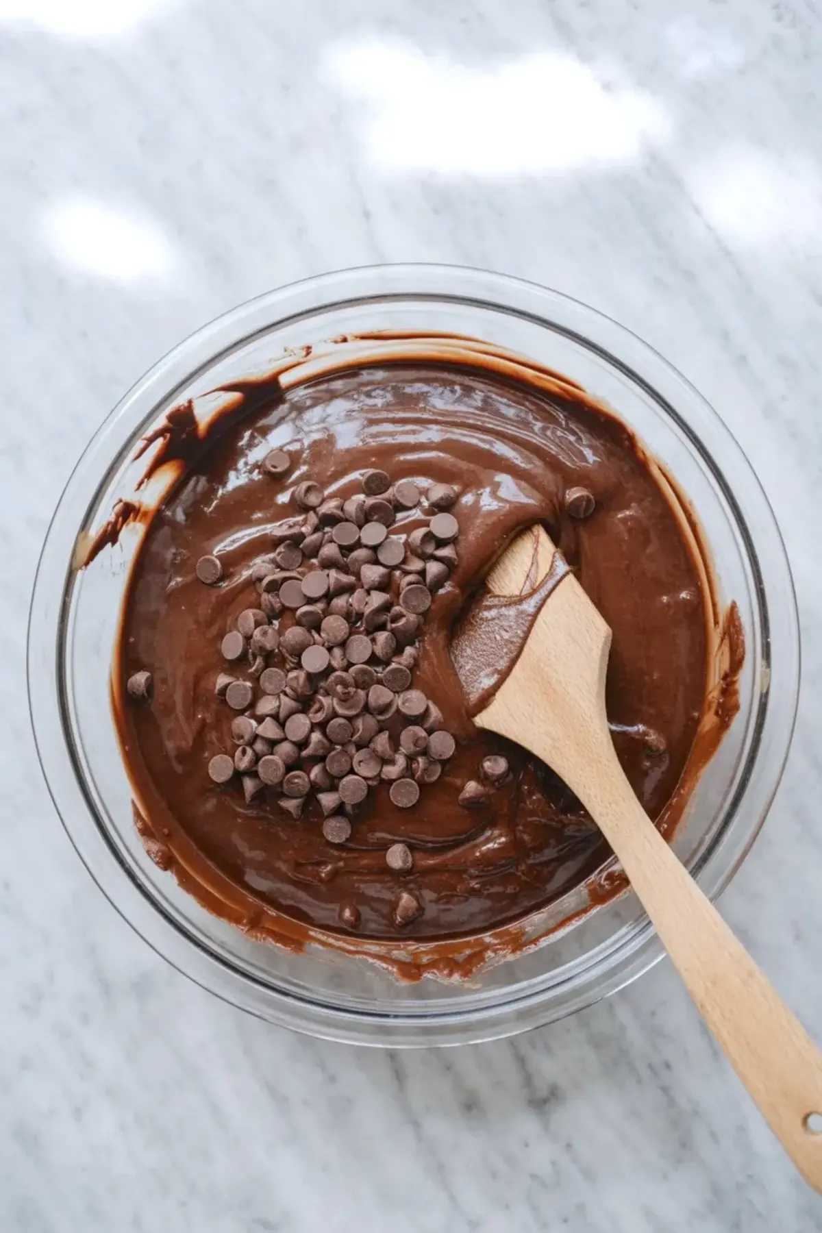 Glass mixing bowl holds thick chocolate brownie batter topped with semi sweet chocolate chips. Wooden spatula stirs glossy dark chocolate mixture on white marble countertop. Homemade brownie recipe preparation shows rich fudgy texture and baking process.